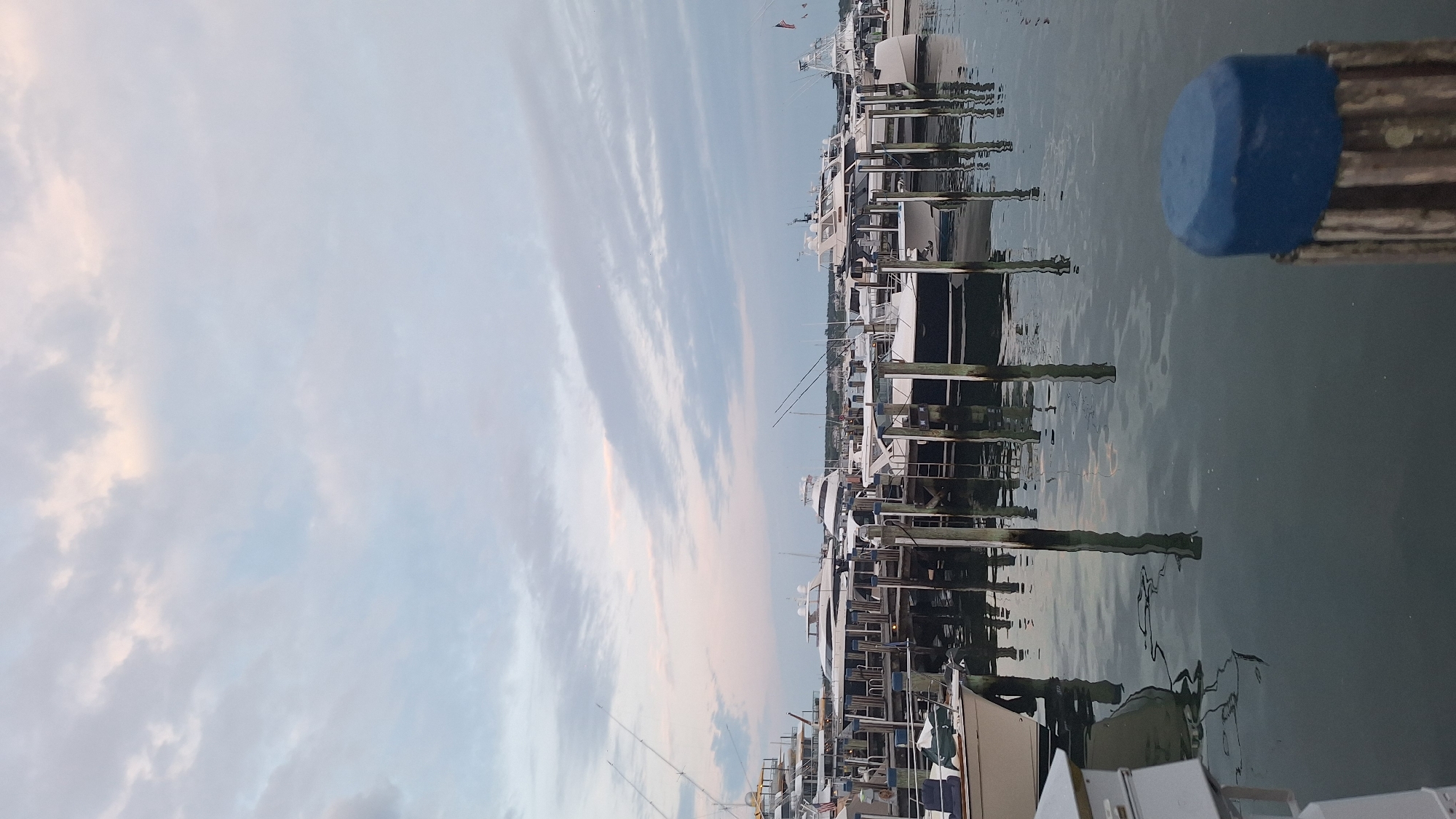 The image shows a view of a marina on Nantucket Island, Massachusetts, United States of America. Several yachts and boats are docked in the water, tied to wooden pilings. The sky is partly cloudy with soft hues of pink and blue, suggesting it is either sunrise or sunset. The water reflects the boats and the sky. In the foreground, a wooden piling with a blue cap is visible. The overall scene depicts a tranquil harbor setting.