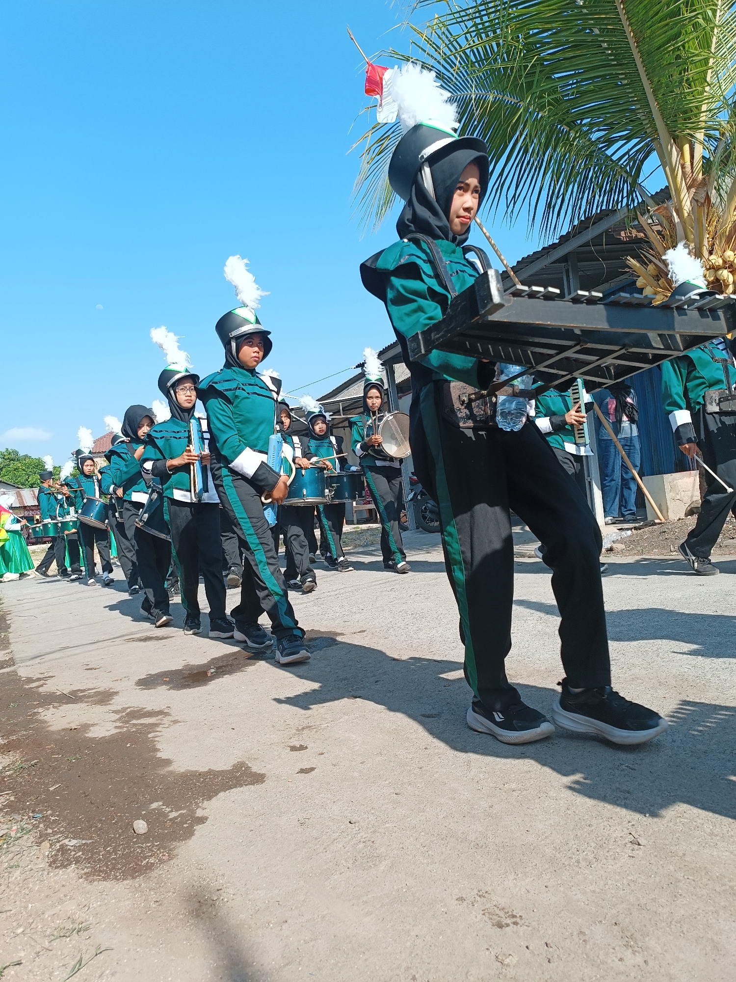 A marching band composed of young women in green and black uniforms with white feather plumes on their helmets walks down a paved road under a clear blue sky. The band members are carrying various instruments, including drums and a xylophone-like instrument held by the lead individual. The scene appears to be in a residential area with houses and palm trees visible in the background. The road is somewhat uneven with patches of dirt and puddles. The overall atmosphere suggests a parade or performance. This event is taking place in Imogiri, Indonesia.