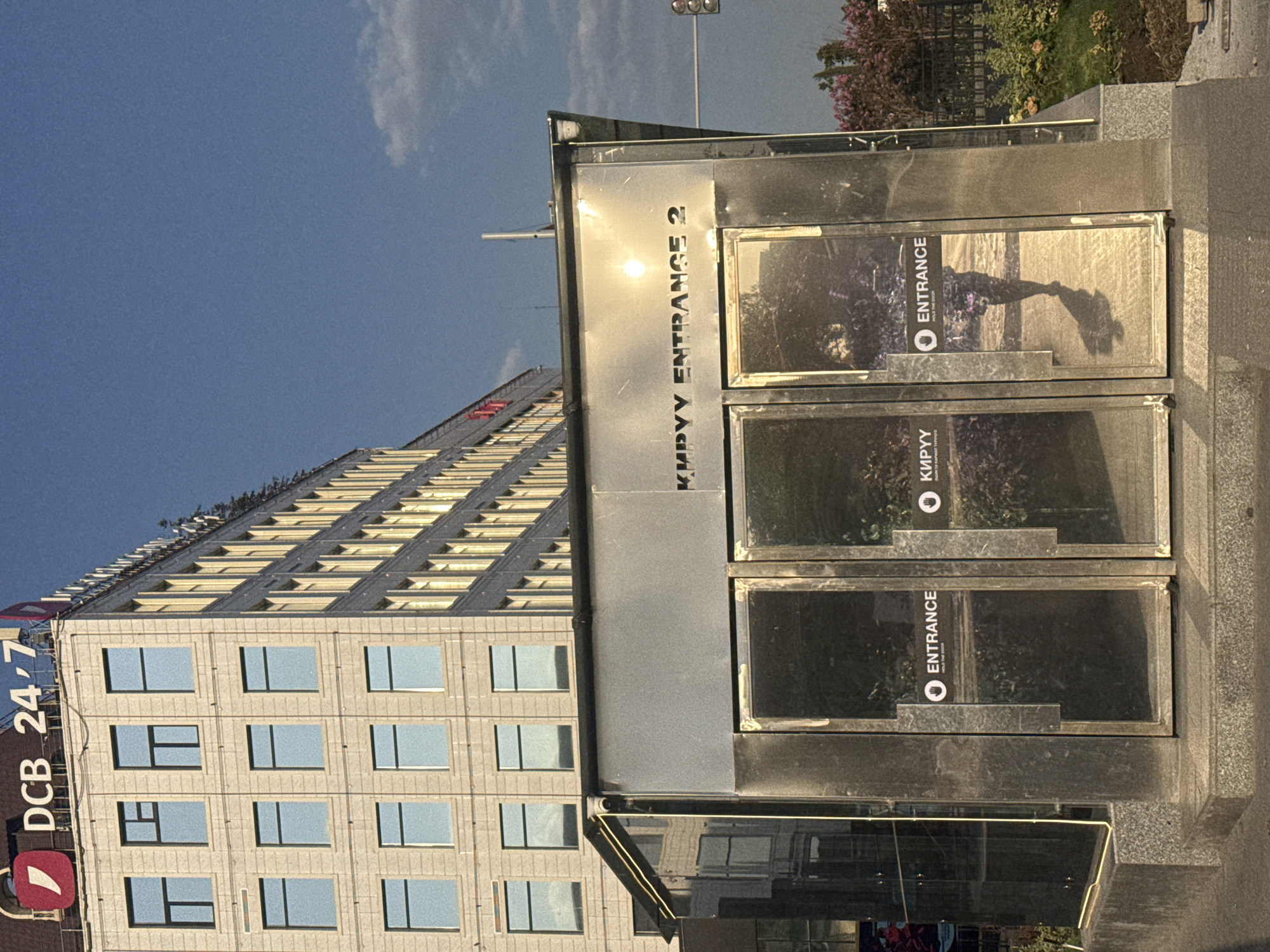 The image shows a modern building in Bishkek, Kyrgyzstan, with a sign that reads "DCB 24/7". In the foreground, there is a metallic structure with three glass doors, two of which have "ENTRANCE" signs. The sun is reflecting off the metallic surface, creating a bright glare. The building behind is made of light-colored stone and has many windows. The sky is blue with some clouds. The overall scene is a street-level view of an urban environment.