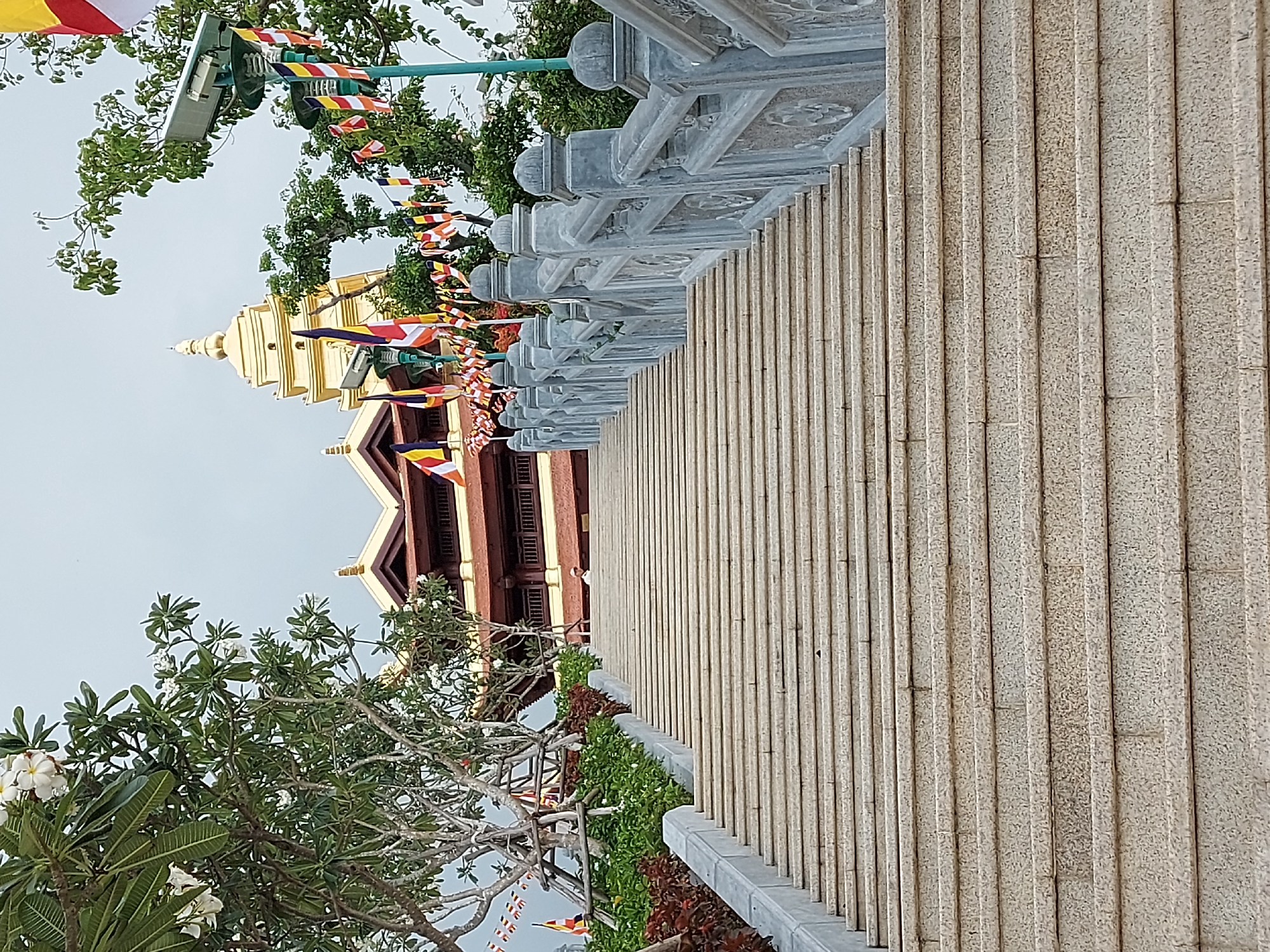 A wide, ornate stone staircase leads upwards towards a temple-like structure with a golden spire. The stairs are flanked by a decorative stone railing with intricate carvings. On either side of the staircase, lush green foliage and trees with white flowers are visible. Colorful prayer flags are strung along the railing and draped from poles, adding a vibrant touch to the scene. In the background, the temple features traditional architecture with a reddish-brown roof and yellow accents. A single person, wearing a white garment, can be seen walking up the stairs. The sky is overcast. The scene is consistent with the entrance to a Buddhist temple, likely in a tropical or subtropical region. This image was taken in Diên Khánh, Vietnam.