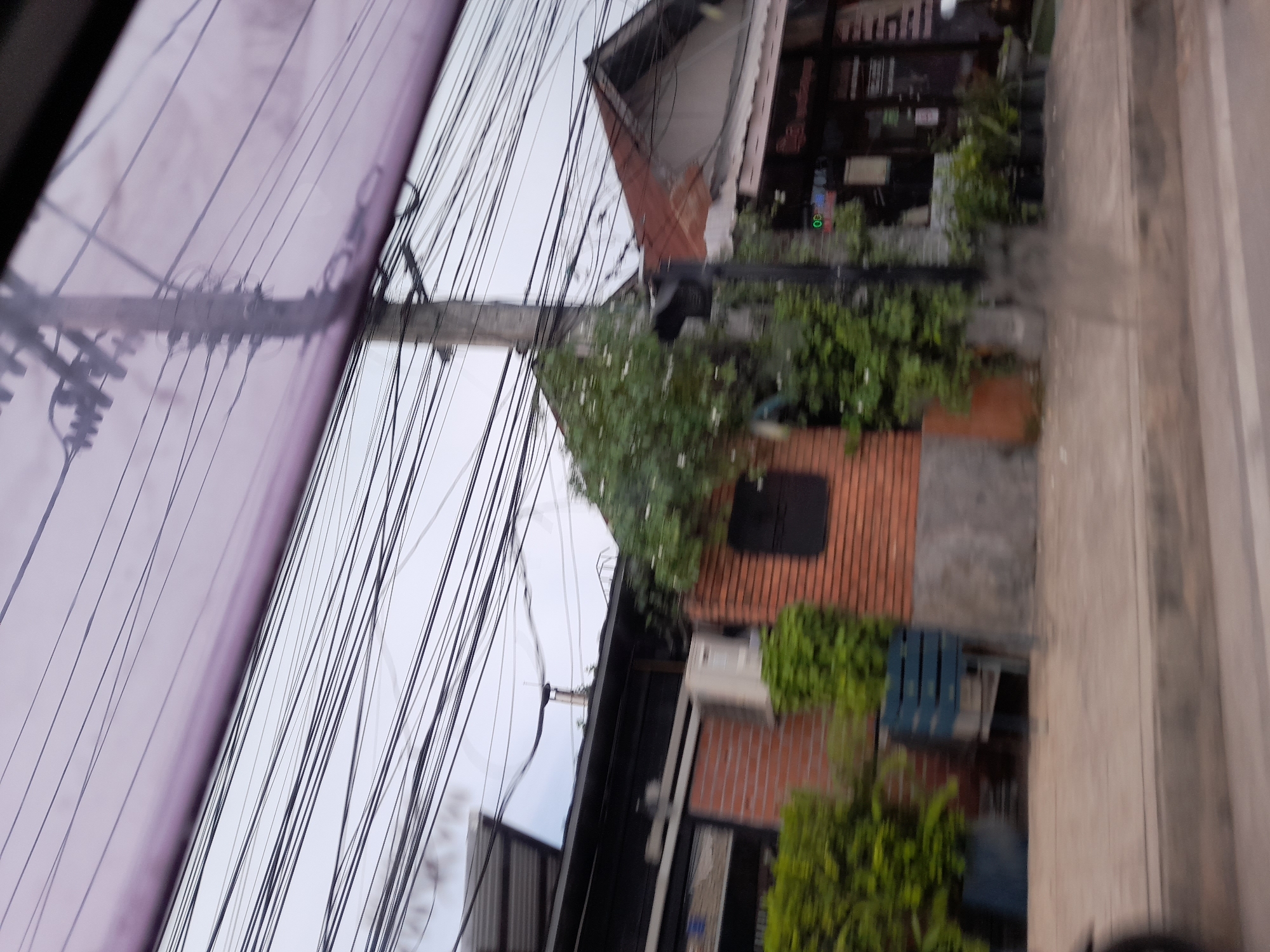 The image captures a street view in Baan Mae Nam, Thailand, characterized by an overcast sky and a dense network of overhead wires. In the foreground, a paved road and sidewalk are visible, with a blurry object, possibly a motorcycle wheel, on the left. Buildings line the sidewalk, one constructed with red brick and featuring large green bushes and a blue slatted bench. Another building to the right has a corrugated metal roof and signage, suggesting a commercial establishment. A traffic light is positioned on a pole amidst lush green foliage, indicating an intersection or a pedestrian crossing. The overall scene conveys a sense of a bustling urban environment with functional infrastructure and natural elements.