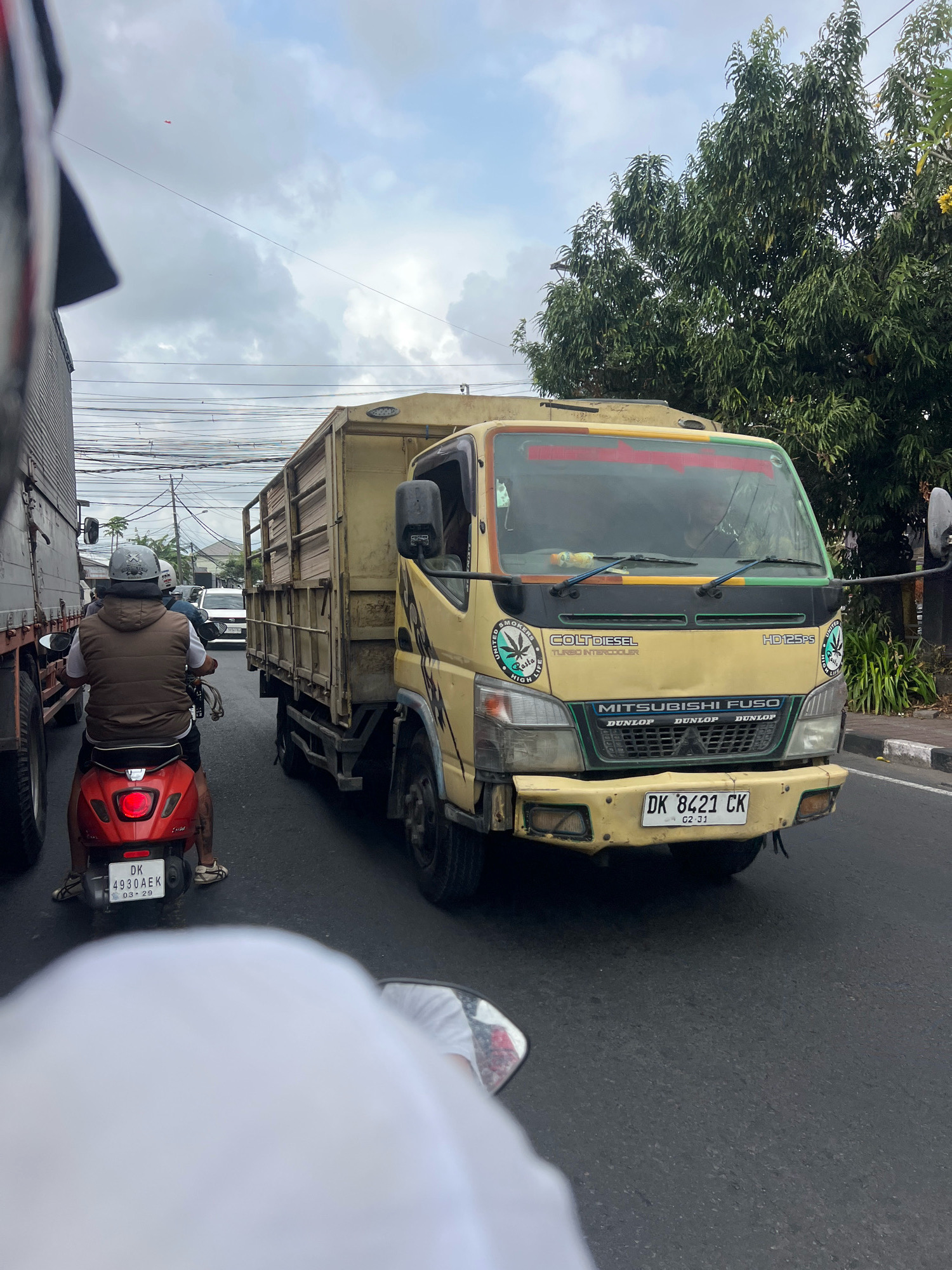 The image captures a view from the perspective of someone riding a red scooter in traffic. Directly in front is a yellow Mitsubishi Fuso truck with a wooden cargo bed. The truck has the license plate "DK 8421 CK". To the left of the truck, another large truck is visible. The scene appears to be on a street in Denpasar, Indonesia, indicated by the "DK" prefix on the license plate, which is specific to Bali. The road is paved, and there are power lines overhead. Lush green trees are on the right side of the road, and a sidewalk with a curb is also visible. The sky is overcast with clouds. The scooter rider in front of the camera is wearing a helmet and a vest over a t-shirt.