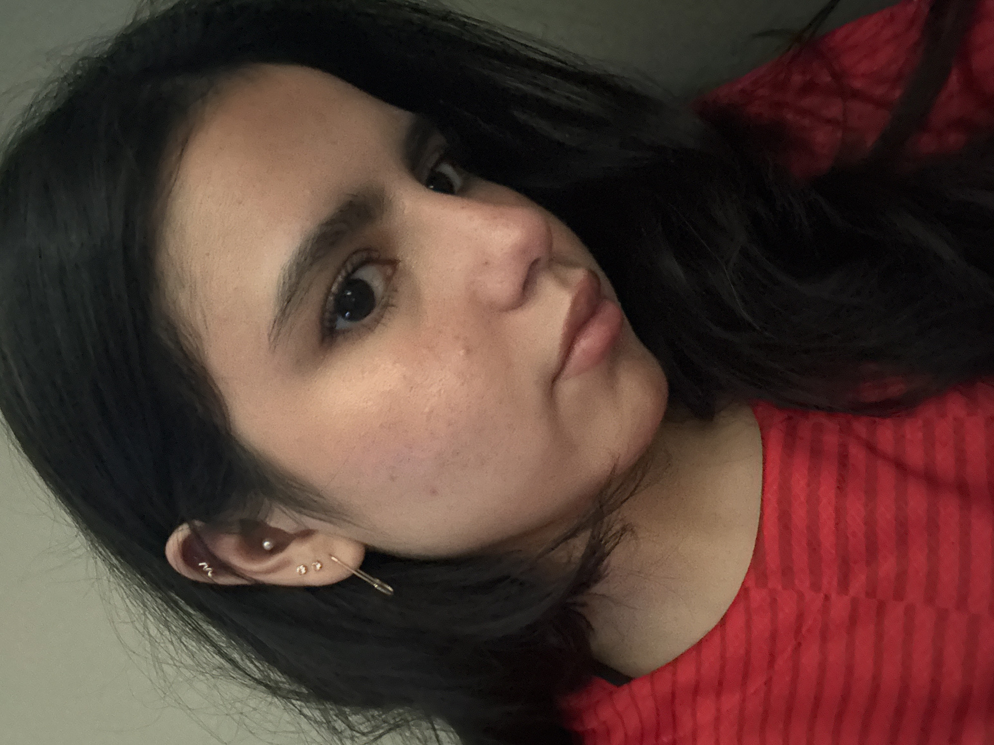 A close-up, angled selfie shows a woman with dark hair, likely in her late teens or early twenties. Her hair is long and loosely styled, falling around her face and shoulder. She is wearing a red shirt with subtle vertical stripes. Her skin tone is fair, with some freckles visible on her cheeks. She has dark eyes and eyebrows. She is wearing multiple earrings in her left ear. The background is slightly out of focus, with what appears to be a light-colored wall. The location, College Station, United States of America, is not evident from the visual information provided in the image.
