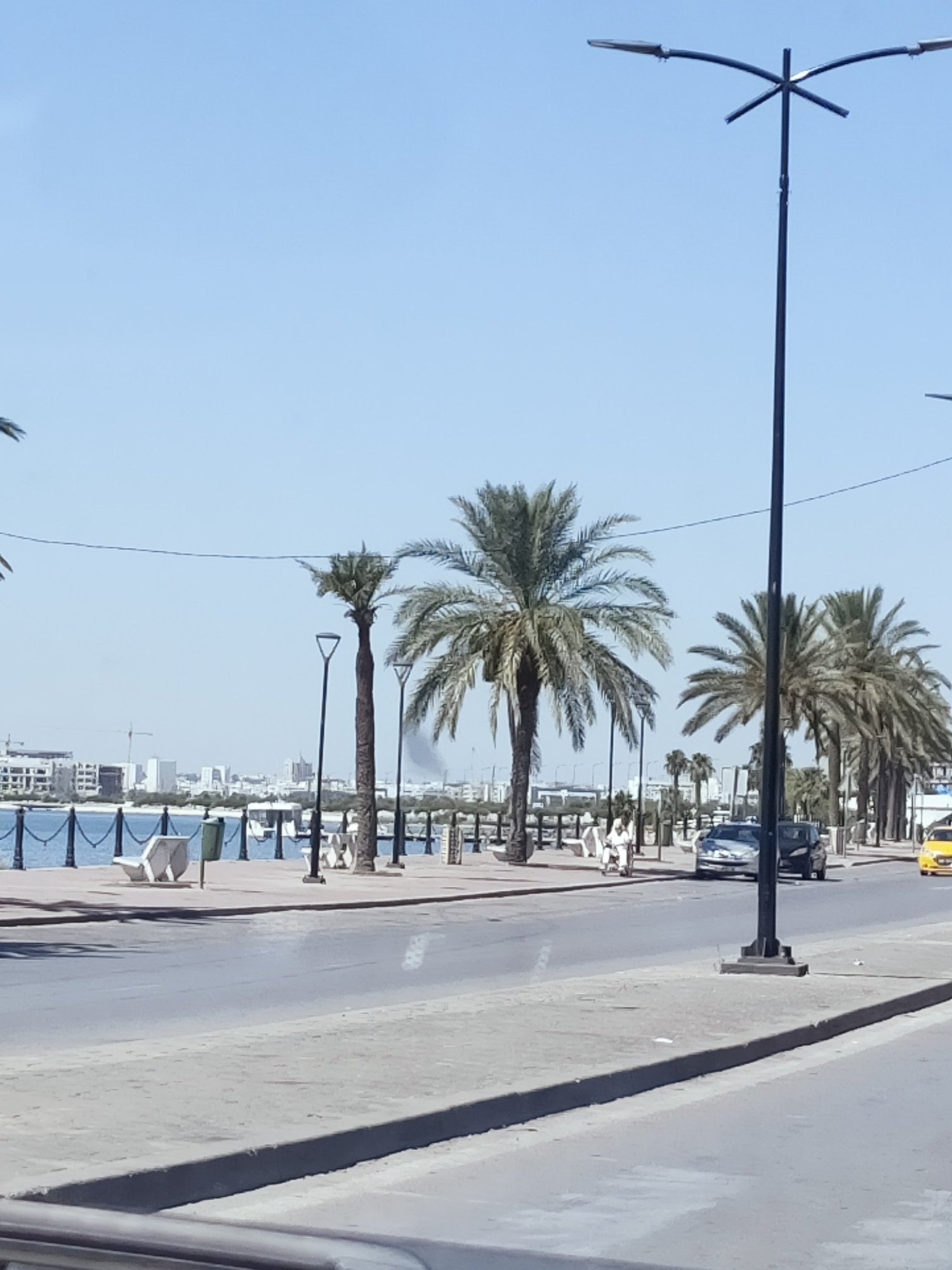 The image depicts a sunny day scene along the Tunis coast, likely near the La Goulette area. In the foreground, a paved road stretches across the frame, with a pedestrian walkway to its left. The walkway features several palm trees lining the waterfront, which is separated by a black metal fence. Beyond the fence, a body of blue water is visible, with a city skyline in the distance, characterized by buildings and construction cranes. Several streetlights with multiple fixtures are positioned along the walkway and road.
On the walkway, two individuals are seen walking, one in white attire. Several cars are present on the road, including a silver sedan and a black car. A yellow taxi is also visible further down the road. The environment suggests a popular urban promenade, potentially a leisure area in Tunis, Tunisia.
