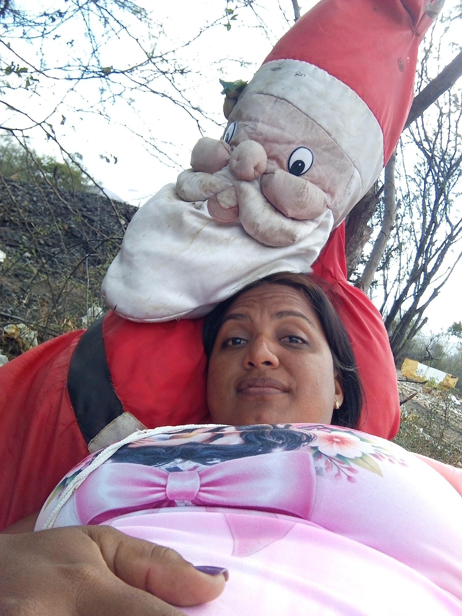 A woman is lying down, her head resting on a large, red and white Santa Claus doll. The woman has dark hair and is wearing a pink shirt with a bow and floral print. Her left hand is visible in the foreground with purple nail polish. The background consists of trees and a somewhat unclear, outdoor environment. The image was taken in Altagracia, Venezuela.