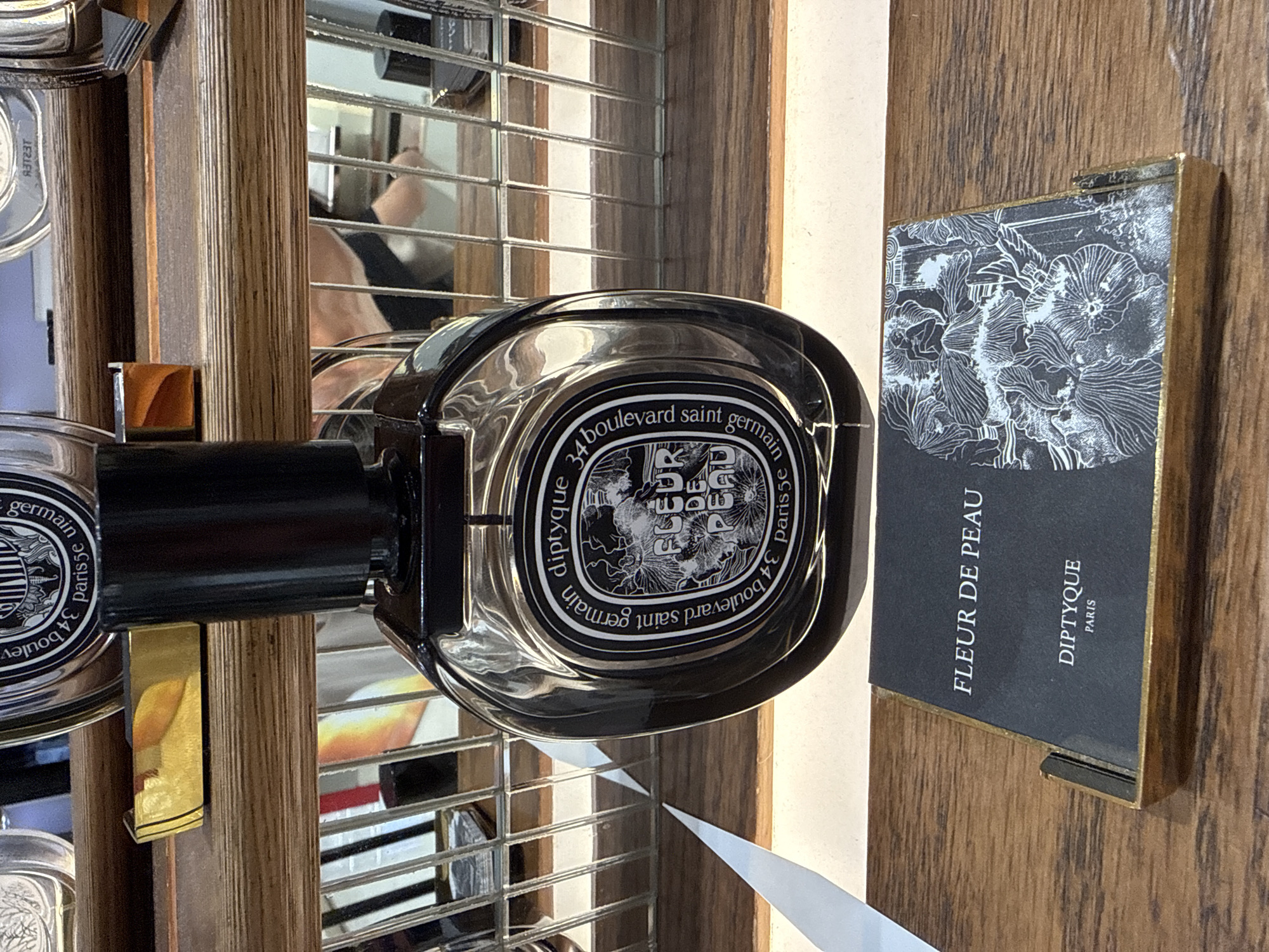The image displays a Diptyque perfume bottle, specifically "Fleur de Peau," positioned on a wooden shelf. The bottle is clear with a black cap and features a circular label with ornate black and white graphics and text. Below the bottle, a small card with the same "Fleur de Peau" and "Diptyque Paris" branding is also visible, nestled in a golden frame. The background shows more shelves with other items and reflections in what appears to be a retail environment. There are no people or specific Kuta, Indonesia landmarks visible in the image.