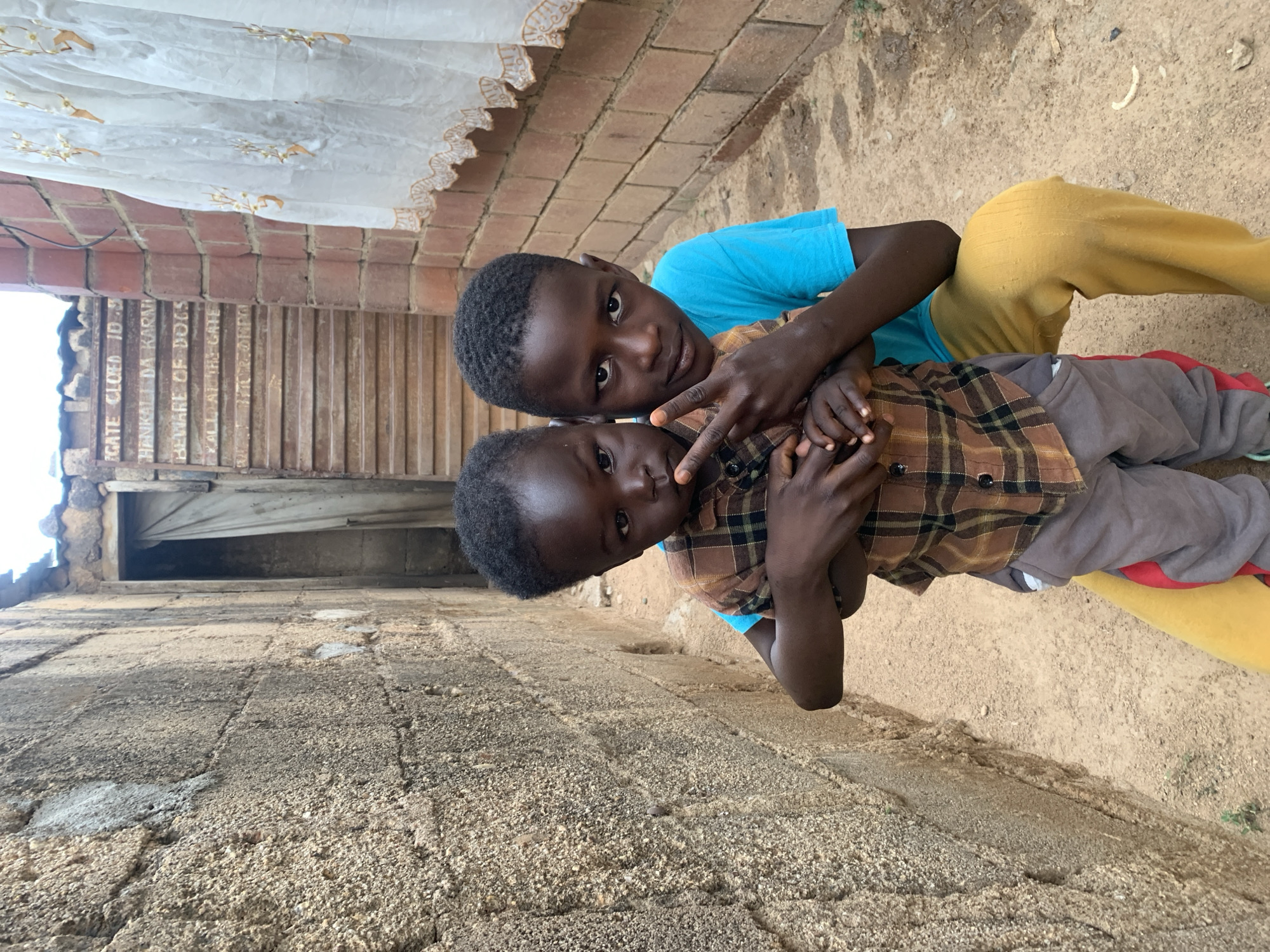 Two young boys are posing for the camera outside a single-story brick building in Jos, Nigeria. One boy is wearing a blue shirt and yellow pants, while the other is wearing a brown and black plaid shirt with gray pants. The boys are holding each other in what seems to be a friendly manner. The building has an overhanging awning with a curtain hanging down. There appears to be an inscription on the building behind the boys. The ground is sandy and there are signs of weathering on the brickwork.