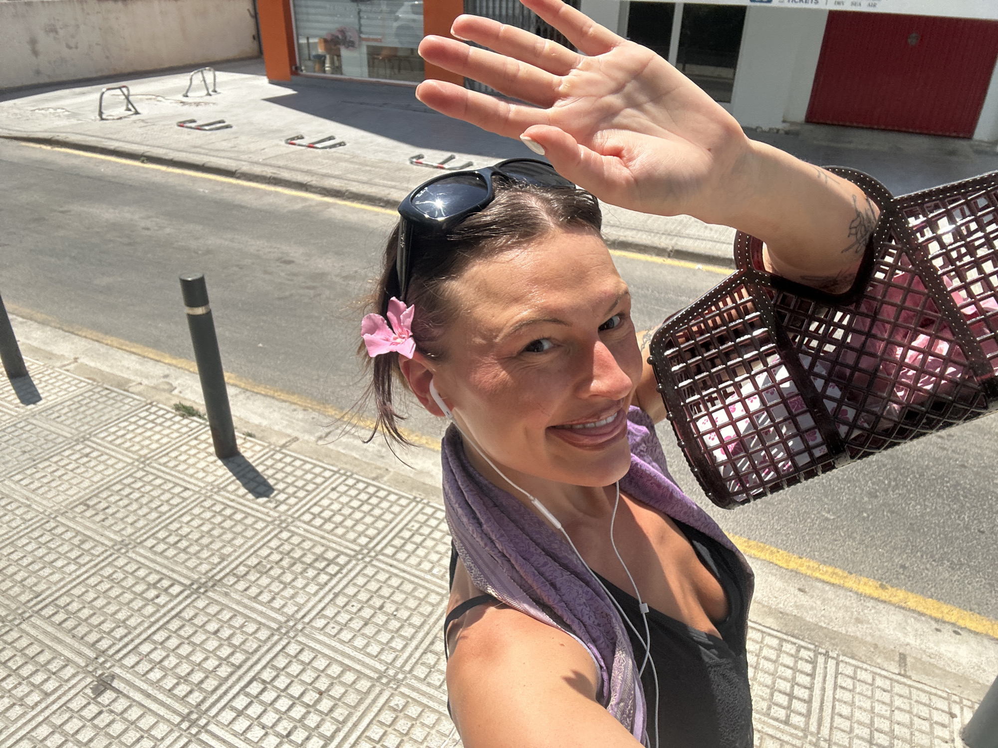 A woman is taking a selfie in the city of Eivissa, Spain. She is wearing a black tank top, a lavender scarf around her neck, and sunglasses on top of her head. A pink flower is tucked behind her ear. She has a hand raised to shield her eyes from the sun. She is carrying a brown woven basket. The background shows a street with metal barriers and the facade of a building.