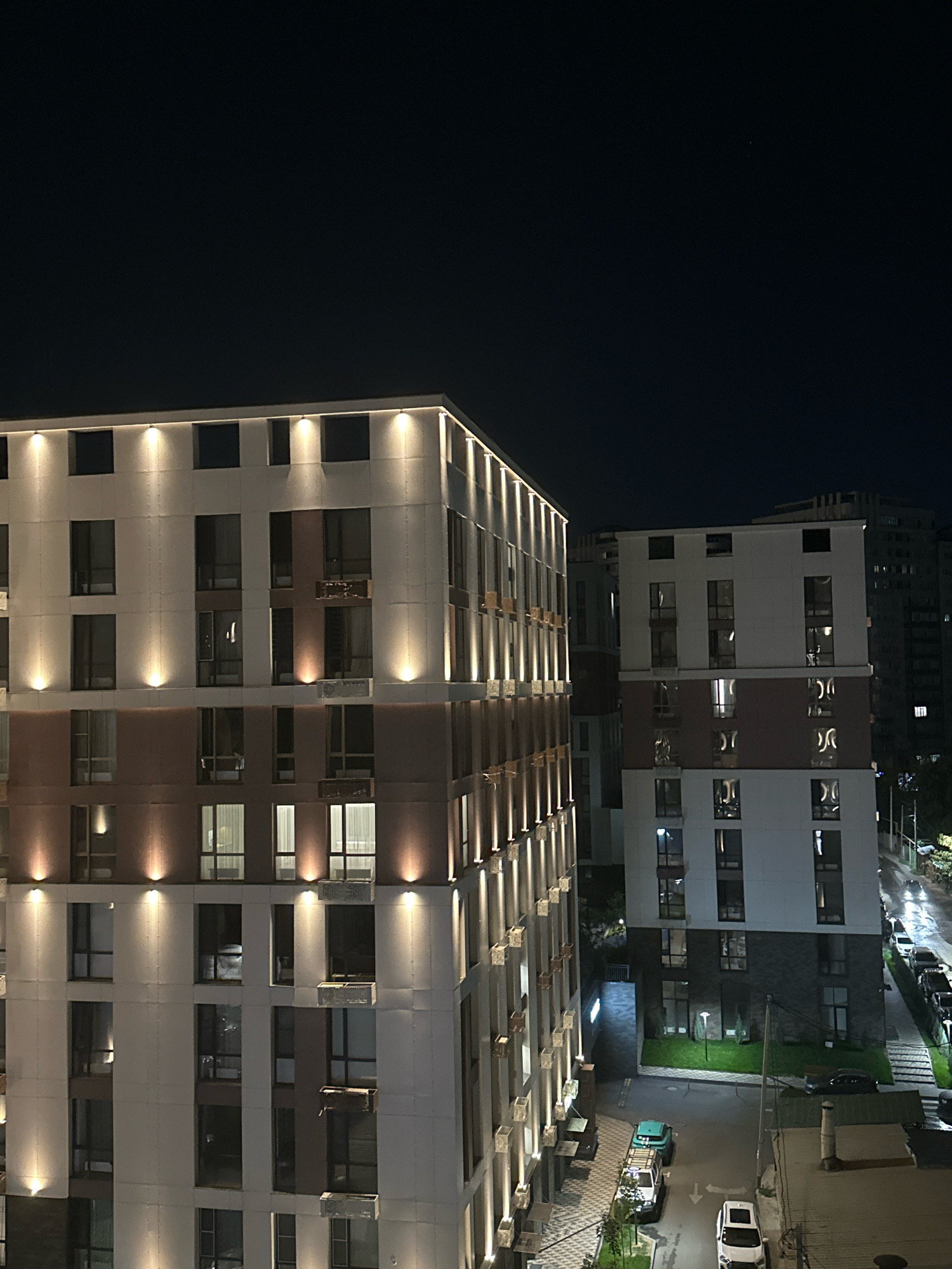 The image shows a night-time exterior view of modern apartment buildings in an urban environment. Two prominent buildings are in the foreground, with their facades illuminated by warm-toned exterior lighting. The building on the left is taller and features a pattern of vertical lights. The building on the right is also multi-story, with a darker lower section and lighter upper sections. Numerous windows are visible on both buildings, some of which show interior lighting.
Below the buildings, a street is visible with parked cars. A white SUV and a turquoise and white utility vehicle are parked on the street. The ground surface is paved, and there are some patches of grass. In the background, other buildings and structures are dimly visible against the dark night sky. The overall impression is of a well-lit residential area at night.