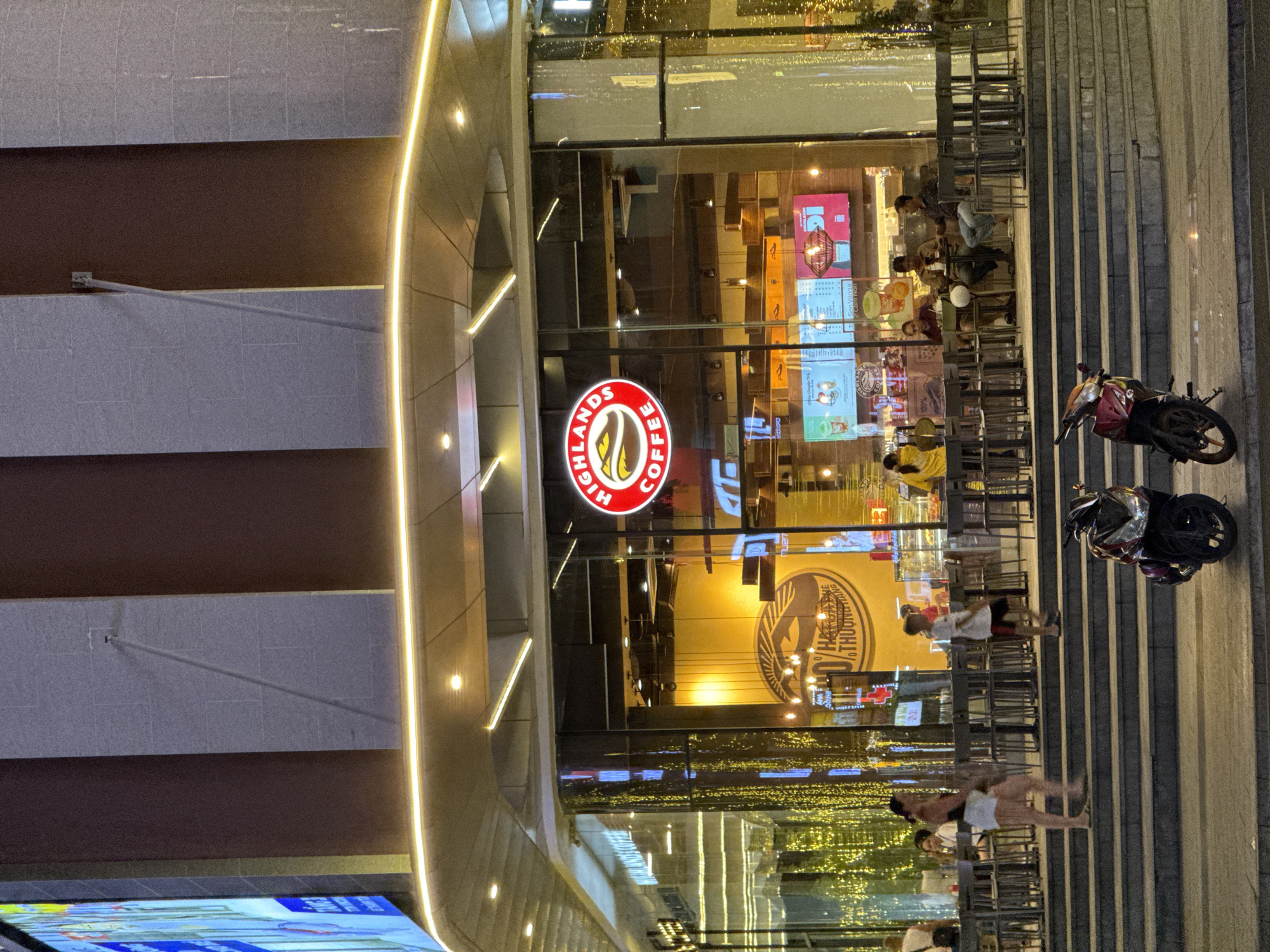 The image depicts a street-level view of a Highlands Coffee shop in Nha Trang, Vietnam, at dusk. The shop features a prominent circular logo with the words "HIGHLANDS COFFEE" and a stylized coffee bean. Large glass windows reveal the interior, where several patrons are seated at tables. The exterior is illuminated by a warm yellow light emanating from within the shop and a strip of yellow lights along the building's facade. A set of concrete stairs leads up to the shop's entrance. Two motorcycles are parked in the foreground on the sidewalk. The overall atmosphere is that of a busy urban evening.