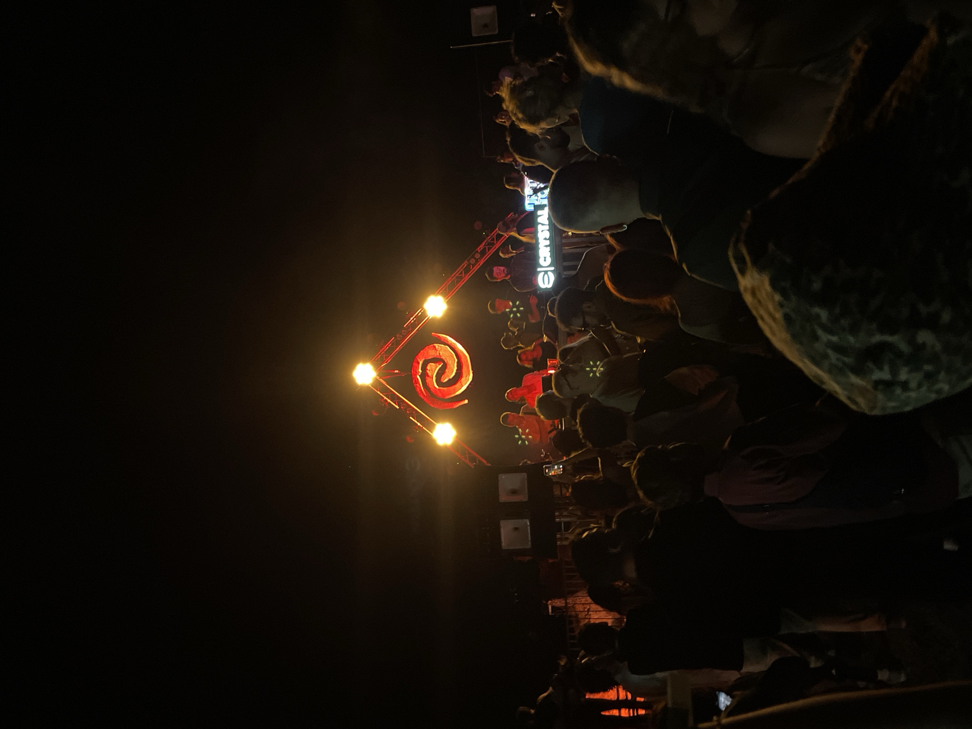 The image depicts a nighttime scene in Budva, Montenegro, likely a concert or outdoor event. A crowd of people is gathered, facing a stage with a red spiral design illuminated by bright lights. Above the stage, a sign with the word "CRYSTAL" is visible. The event appears to be taking place outdoors, with darkness surrounding the illuminated stage area. People in the crowd are seen taking pictures with their phones.