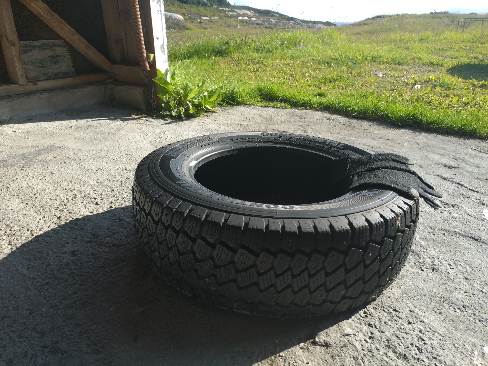 An outdoor, slightly angled, eye-level medium shot shows a car tire lying on a concrete surface in Averøy, Norway. A black glove is draped over the top of the tire. The tire, with its tread and metal studs visible, casts a shadow to its left. Behind the tire, a section of a wooden structure with exposed beams and corrugated metal is visible. Beyond that, green grass, some rocks, and a body of water under a bright sky complete the background. The scene appears to be a sunny day, with strong shadows indicating the sun's position.