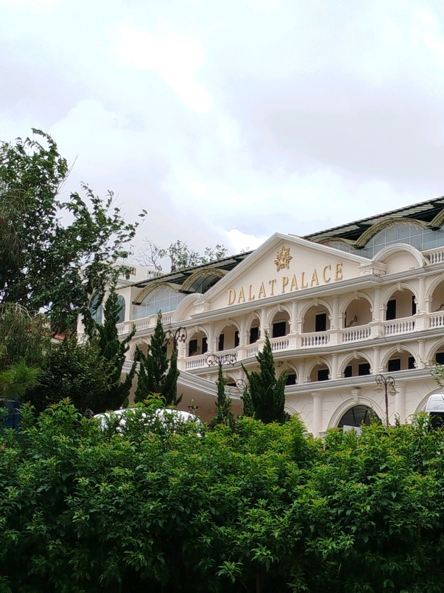 The image shows the exterior of the Dalat Palace Hotel, a historic landmark in Da Lat, Vietnam. The hotel is a grand, cream-colored building with a distinctive architectural style featuring multiple arches, balconies, and ornate detailing. The name "DALAT PALACE" is prominently displayed in gold lettering above the main entrance. Green trees and lush foliage are in the foreground, partially obscuring the lower portion of the building. The sky above is overcast with clouds. The scene appears to be captured on a daytime with soft, diffused light.