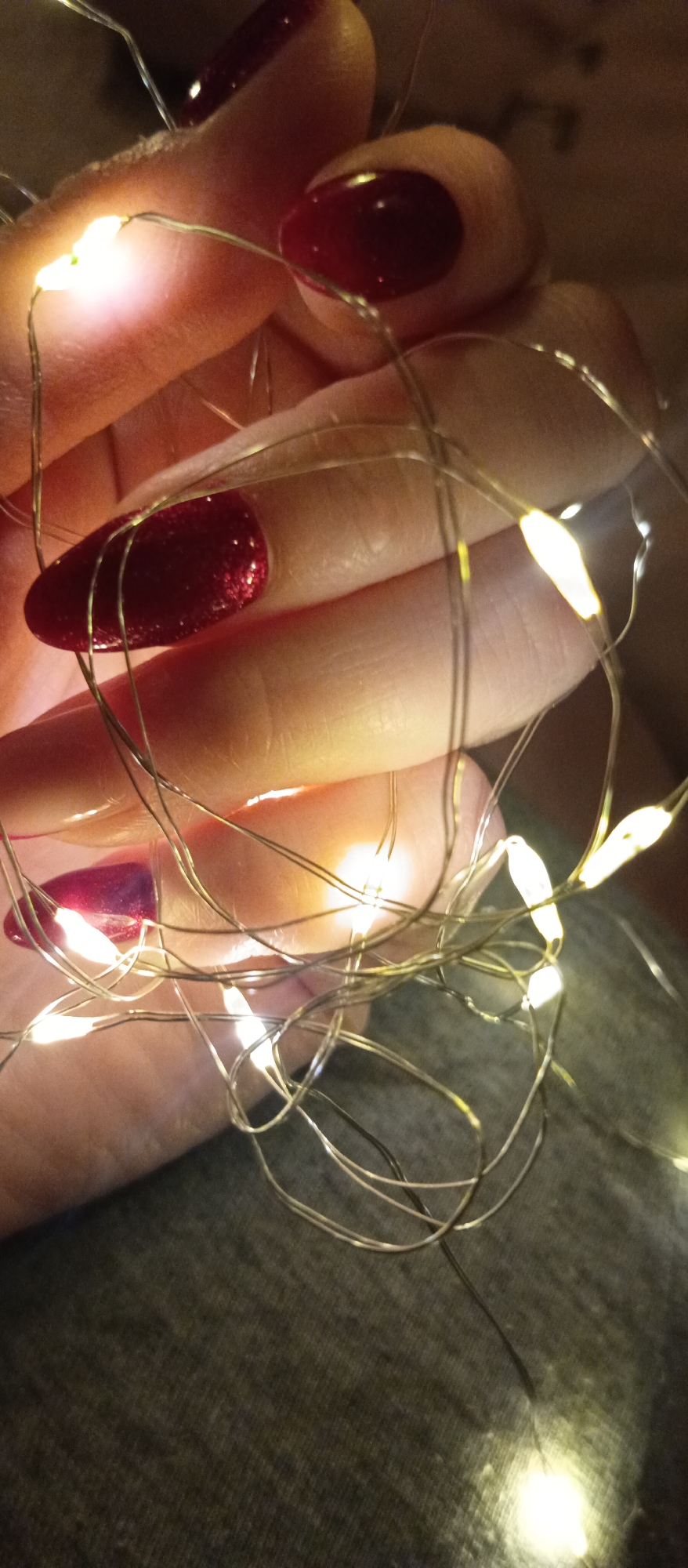 A close-up shot depicts a hand with manicured nails, holding a tangle of string lights. The nails are painted with a dark red, possibly glittered, polish. The fingers are slightly curled and appear relaxed. The string lights are silver-colored and interspersed with small, warm white LED bulbs. The background is out of focus and appears to be a gray fabric, likely clothing. The location and city are unknown.