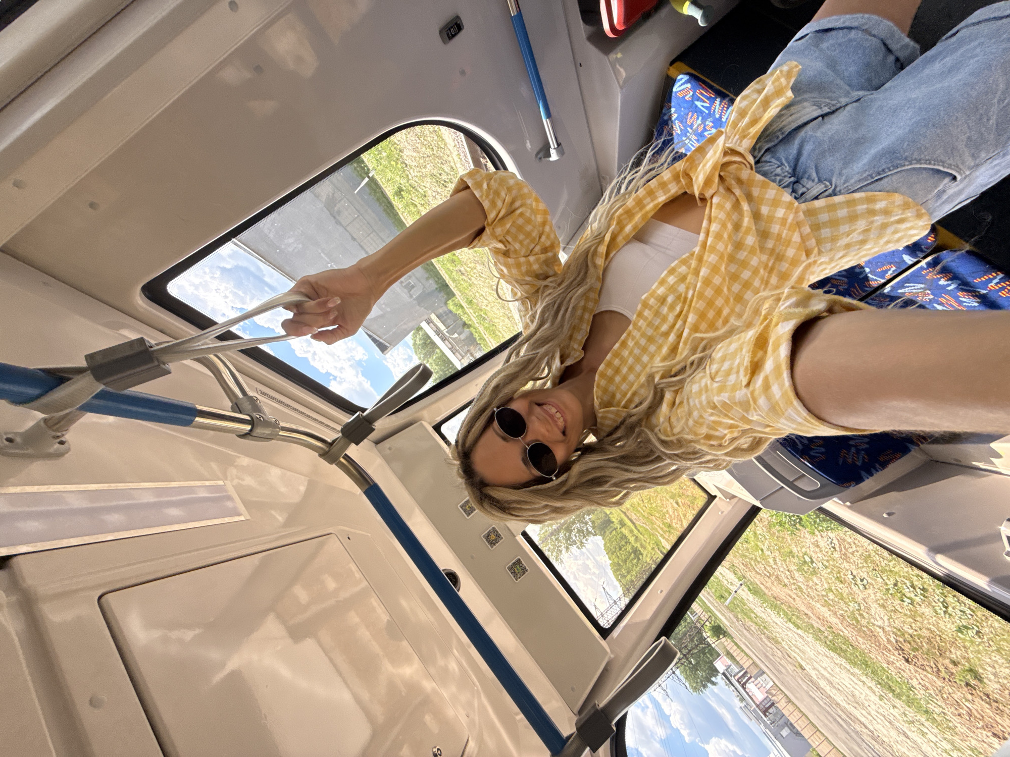A young woman is taking a selfie while riding on public transportation, possibly a bus, in Заволжье, Russia. She is blonde, wearing sunglasses, a white top, and a yellow checkered shirt tied around her waist. She is holding onto a handrail above. The background shows scenery visible through the bus window, which includes green fields and a partially visible building. The interior of the bus is clean, with white panels, blue handrails, and patterned seat covers.