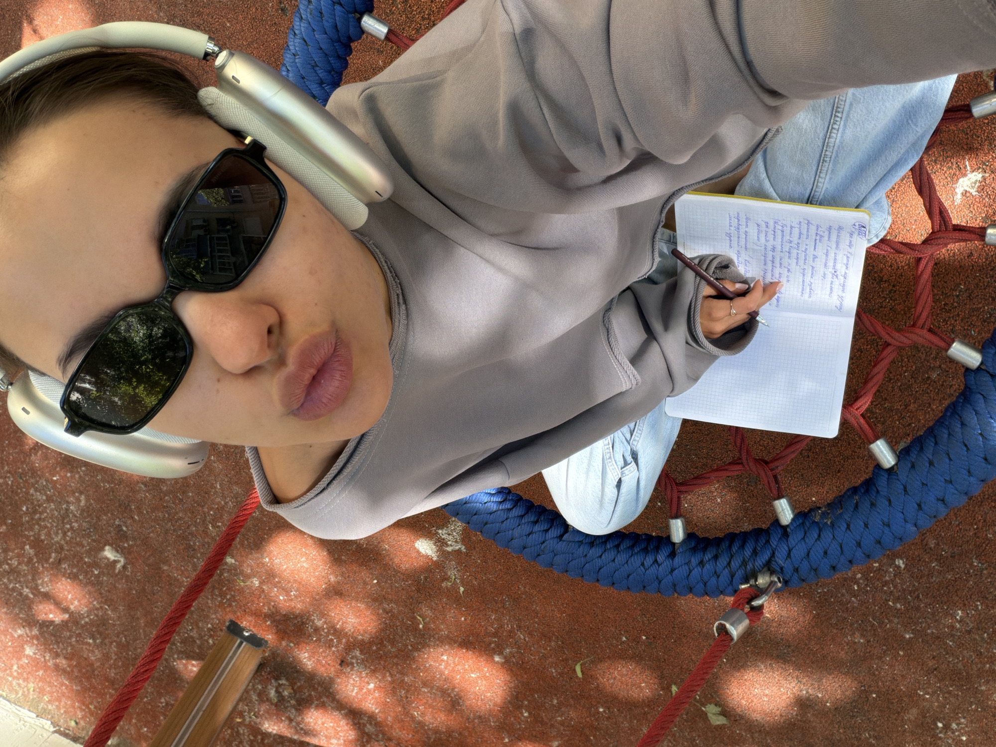 A young adult female is captured in a high-angle selfie, seated on a circular net swing within an outdoor playground environment. She wears black rectangular sunglasses and light-colored over-ear headphones, resembling Apple AirPods Max. Her attire consists of a grey long-sleeved sweatshirt and light blue denim jeans. The subject is posing with pursed lips. Her right hand, adorned with a ring on the ring finger and manicured nails, holds a dark pen. She is writing in an open, grid-lined notebook with a yellow edge, where Cyrillic script is visible. The swing features a thick blue rope perimeter and a red rope net interior, connected by silver metal fixtures. The ground beneath is a reddish-brown rubberized safety surface, marked with dappled sunlight. Reflections in the sunglasses reveal multi-story urban buildings and green foliage, consistent with a city setting. The presence of Cyrillic script strongly indicates the scene is located in an urban area within Moscow, Russia.
