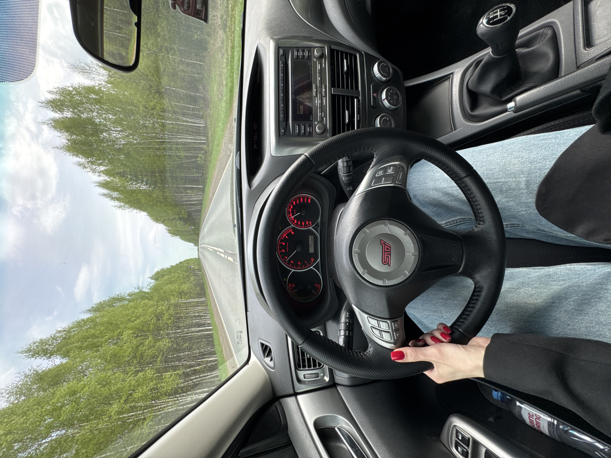 The image captures a driver's perspective inside a Subaru WRX STI, facing forward on a road with birch trees lining each side. The dashboard includes a factory head unit with a CD/AUX player, climate control knobs, and gauges showing the car has 175,980 kilometers on it. The steering wheel features the STI logo. The driver's hand, with red nail polish, is visible on the steering wheel. The gear shifter indicates a manual transmission. A glimpse of the exterior through the windshield shows a straight road with a white line. A water bottle is in the door. The image could be in or near Kamennyi Kliuch, Russia.