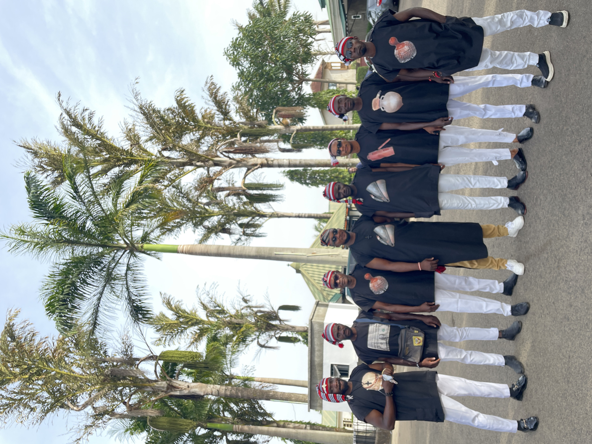 A group of seven men stand in a vertical line on a paved surface, possibly a street or driveway. The location appears to be in Jos, Nigeria, based on the cultural attire of the men. Each man wears a black short-sleeved tunic with a distinct graphic on the front, white trousers, and a traditional striped hat. The men are arranged from the bottom to the top of the frame and appear to be posing for a photograph.
Behind the men are several tall palm trees and other trees, suggesting an outdoor setting with tropical vegetation. The background also shows a building with a light-colored roof and walls. The sky is light and mostly clear.