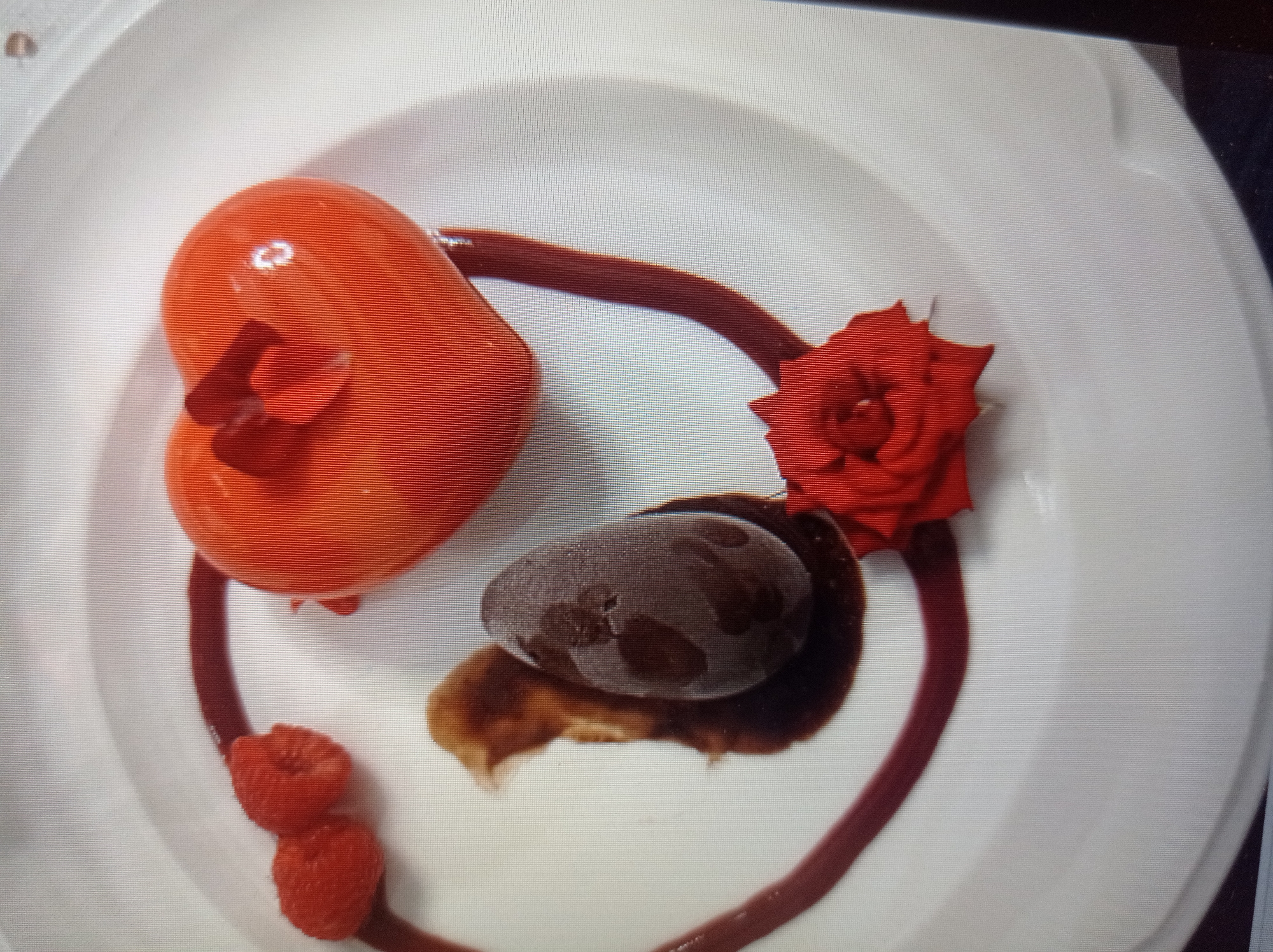 A heart-shaped, orange-glazed dessert sits on a white plate, garnished with two raspberries and a red flower. Beside it is a dark, egg-shaped item in a pool of dark sauce. A thin, dark red drizzle encircles the dessert. The setting is a white plate on a flat surface, with no visible elements of Mukachevo, Ukraine. The scene is a close-up, static shot of the dessert.
