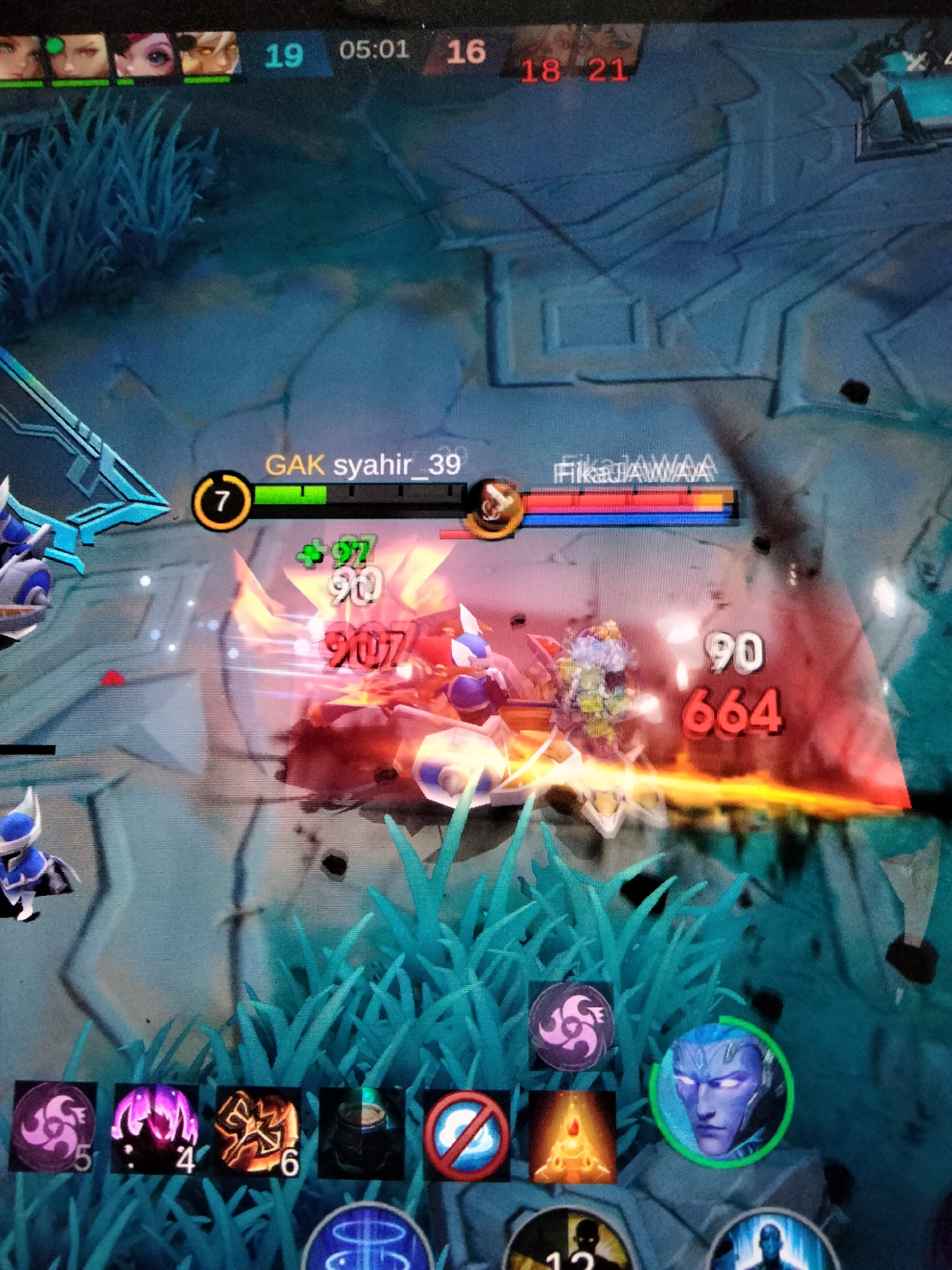This is a screenshot from a mobile game, likely a multiplayer online battle arena (MOBA) game. The scene depicts a battle in progress with two opposing teams.
The foreground shows several game elements:
- **Characters:** Two characters are engaged in combat. One appears to be a blue-skinned humanoid with glowing eyes, possibly a hero character. Another character, partially obscured by the combat effects, seems to be a creature with metallic armor and wings.
- **Damage Numbers:** Large numbers indicating damage dealt are visible, such as "907" and "664," along with smaller numbers like "90" and "+97."
- **Abilities Icons:** At the bottom of the screen, there are icons representing character abilities, some with associated numbers indicating cooldowns or charges (e.g., "5," "4," "6").
- **UI Elements:** A player's name, "GAK syahir_39," is displayed above a health bar. Another player's name, "FikaAWAA," is also visible. A timer at the top shows "05:01," indicating the game's duration. Scores for each team are also present: "19" for one team and "16" for the other, with "18 21" also displayed, possibly representing player kills.
The background depicts a fantasy-themed environment with rocky terrain and stylized blue foliage. The overall lighting suggests it is an in-game environment, not a real-world setting. The emotions conveyed are those of intense action and conflict, common in MOBA games. The presence of player names and team scores suggests a competitive multiplayer match. The context of Payagaboh, Indonesia, suggests this is a screenshot from a player in that region.