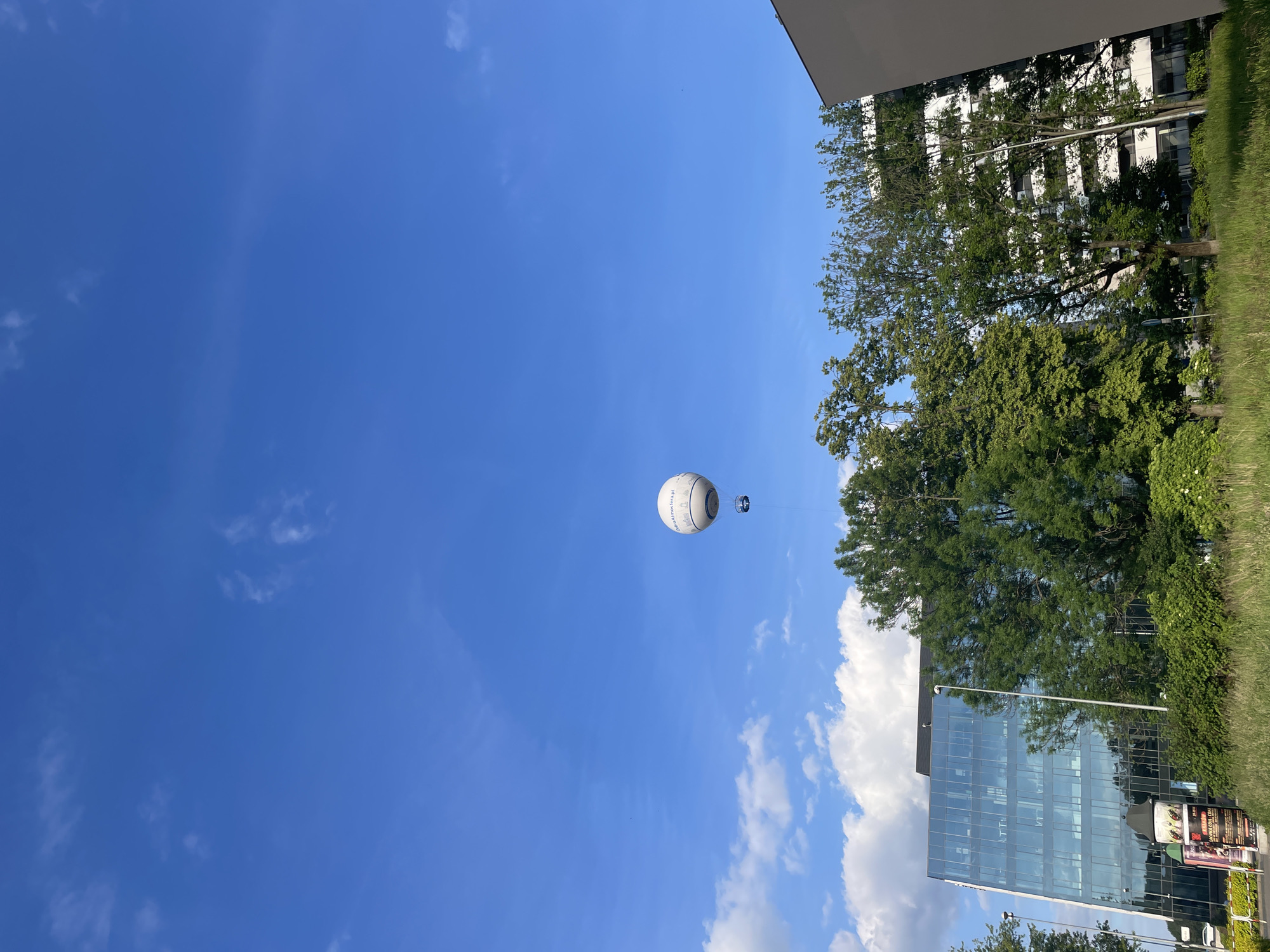 The image shows a white, spherical tethered balloon in a blue sky with scattered clouds. The balloon has text on its side, partially visible as "atmosfera.pl". A small observation basket is suspended below the balloon. The foreground includes trees and buildings, one with a glass facade reflecting the sky. The image's orientation is rotated ninety degrees clockwise. The city is Unknown, Unknown.