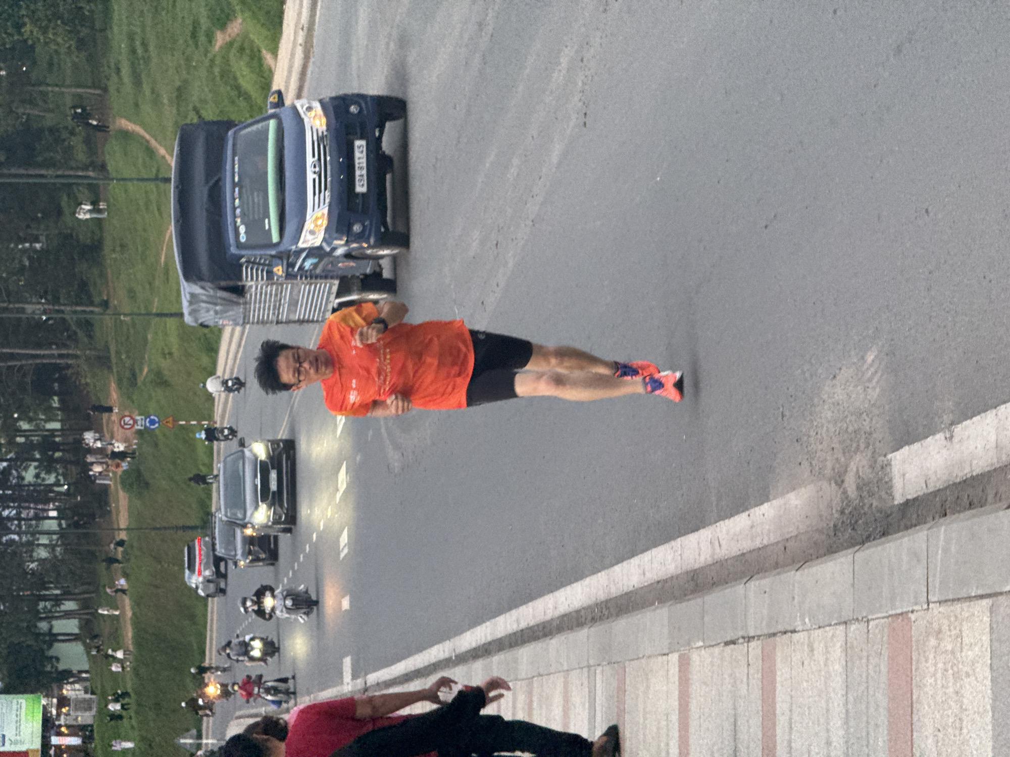 The image shows a man running on a paved road in Da Lat, Vietnam, with a truck and several motorcycles in the background. The man, wearing an orange shirt and black shorts, is in motion, his arms bent and his legs in a running stride. His shoes are bright pink and blue. A blue truck with a tarp-covered bed is driving on the road to the man's right. Several motorcycles are also visible on the road, some with riders. In the background, there are trees and a grassy hillside, suggesting an outdoor, possibly park-like setting within the city. A sidewalk runs along the left side of the road, with some people walking on it. The overall scene depicts a lively urban environment with ongoing activity.