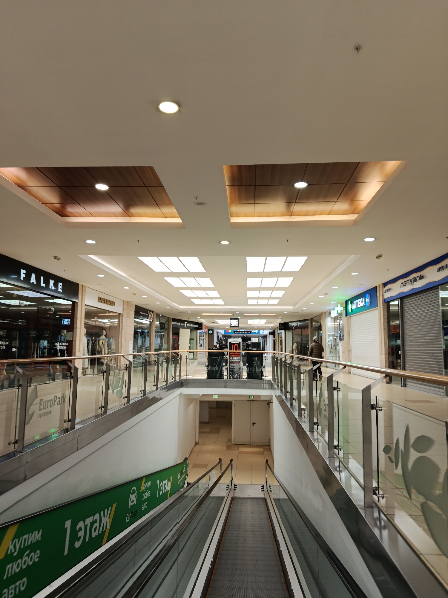 The image shows the interior of a shopping mall, likely in Moscow, Russia, given the Cyrillic text and architectural style. The perspective is from an elevated point, looking down at an escalator. The escalator is marked with a green sign, "1 этаж" and "купим любое авто," suggesting "1st floor" and "we buy any car." Above the escalator is a pedestrian corridor with glass railings.
Several storefronts are visible in the background, including "FALKE," "RAFINAD", АПТЕКА (Pharmacy) and ЛЭТУАЛЬ(cosmetics store). The mall has a modern design with recessed lighting and a mixture of natural and artificial light. A single person can be seen walking in the pedestrian corridor.