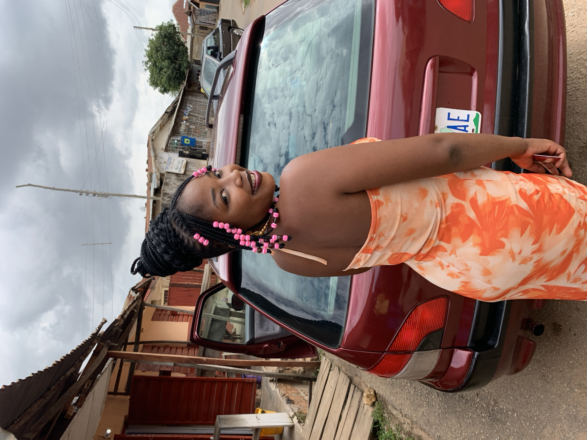 A young adult female with dark skin, styled black braided hair adorned with numerous pink and dark beads, and wearing an orange and white tie-dye strapless dress, stands with her back partially to the camera, smiling over her left shoulder. She is positioned next to the rear of a dark red station wagon, which has its driver's side rear door open. The car's license plate is partially visible, displaying "AE". The background, consistent with an urban street in Jos, Nigeria, features single-story buildings with corrugated metal roofs and red-painted wooden shutters. Utility poles and overhead wires are present, and a dark sedan is parked further down the unpaved road. The sky is overcast and cloudy, with reflections visible in the car's rear window.
