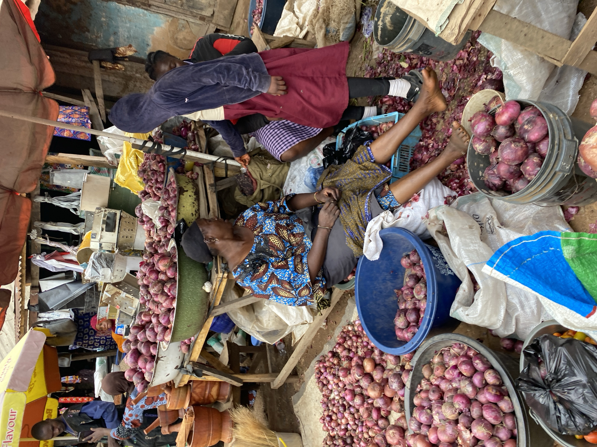 A crowded outdoor market scene in Bukuru, Nigeria features numerous piles of red onions, some in large containers and others spread across the ground. A woman in a vibrantly patterned blue and gold dress and a dark headscarf sits on the ground, her legs crossed, surrounded by onions and a blue plastic basket. To her right, a person wearing a dark blue hooded jacket and a burgundy skirt stands, interacting with someone behind them who is partially visible. In the background, more market stalls are visible with various goods, including clay pots and other produce. The ground is covered in dirt and scattered onion skins. The overall impression is of a busy, bustling marketplace.