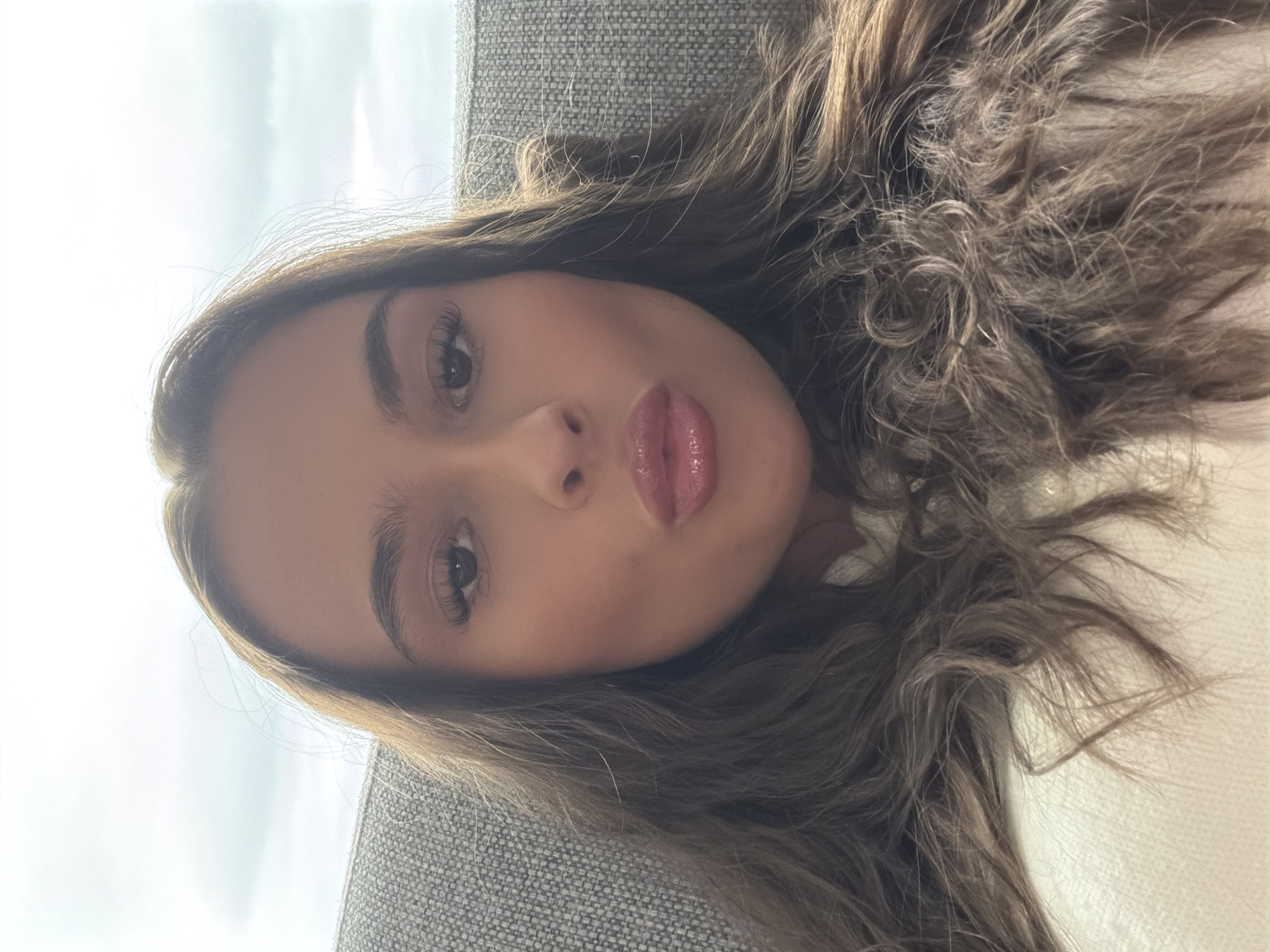 A close-up selfie of a young woman with long, wavy brown hair and fair skin. She is wearing a light-colored top and has her lips pursed. Her eyes are dark brown and she has full lips with lip gloss. She is sitting on a gray sofa against a backdrop of a cloudy sky. The scene is indoors, likely in a residential setting. There is no identifiable information in the image that places it in Harstad, Norway.