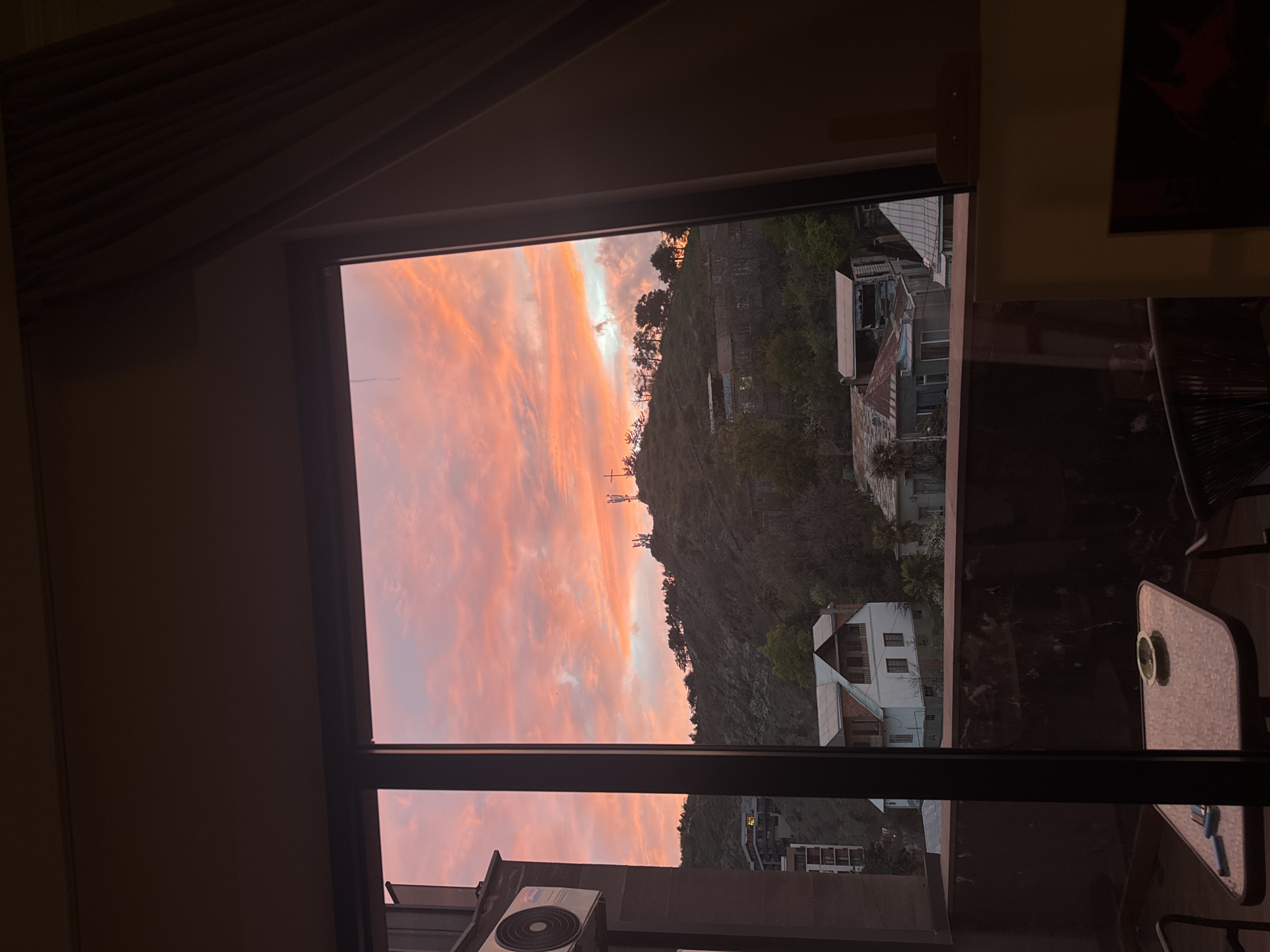 The image displays a view from inside a dark room looking out a large window at what appears to be a sunset or sunrise. The sky is a prominent feature, filled with dramatic clouds colored in vivid shades of orange, pink, and purple, with hints of pale blue and white where the light is brightest.
Beyond the window, a steep, tree-covered mountain or hillside rises, with its dark silhouette contrasting against the colorful sky. Numerous buildings, varying in size and style from multi-story residential structures with white walls to more rustic-looking dwellings with corrugated roofs, are visible scattered across the hillside and at its base. On the distant peak of the mountain, a distinct cross-like structure or antenna stands tall against the sky.
The interior of the room is dark, showing parts of the window frame and what seems to be a ceiling or wall structure. Reflections on the window reveal elements from the room or an adjacent balcony: a small, light-colored outdoor table with a circular object on it (possibly a cup or ashtray), and part of a dark, woven chair. An outdoor air conditioning unit is mounted on a structure below the window on the lower left, with the brand name "Midea" visible. No people are present in the image. The time of day is clearly either dawn or dusk, given the vibrant sky colors and the dark interior.