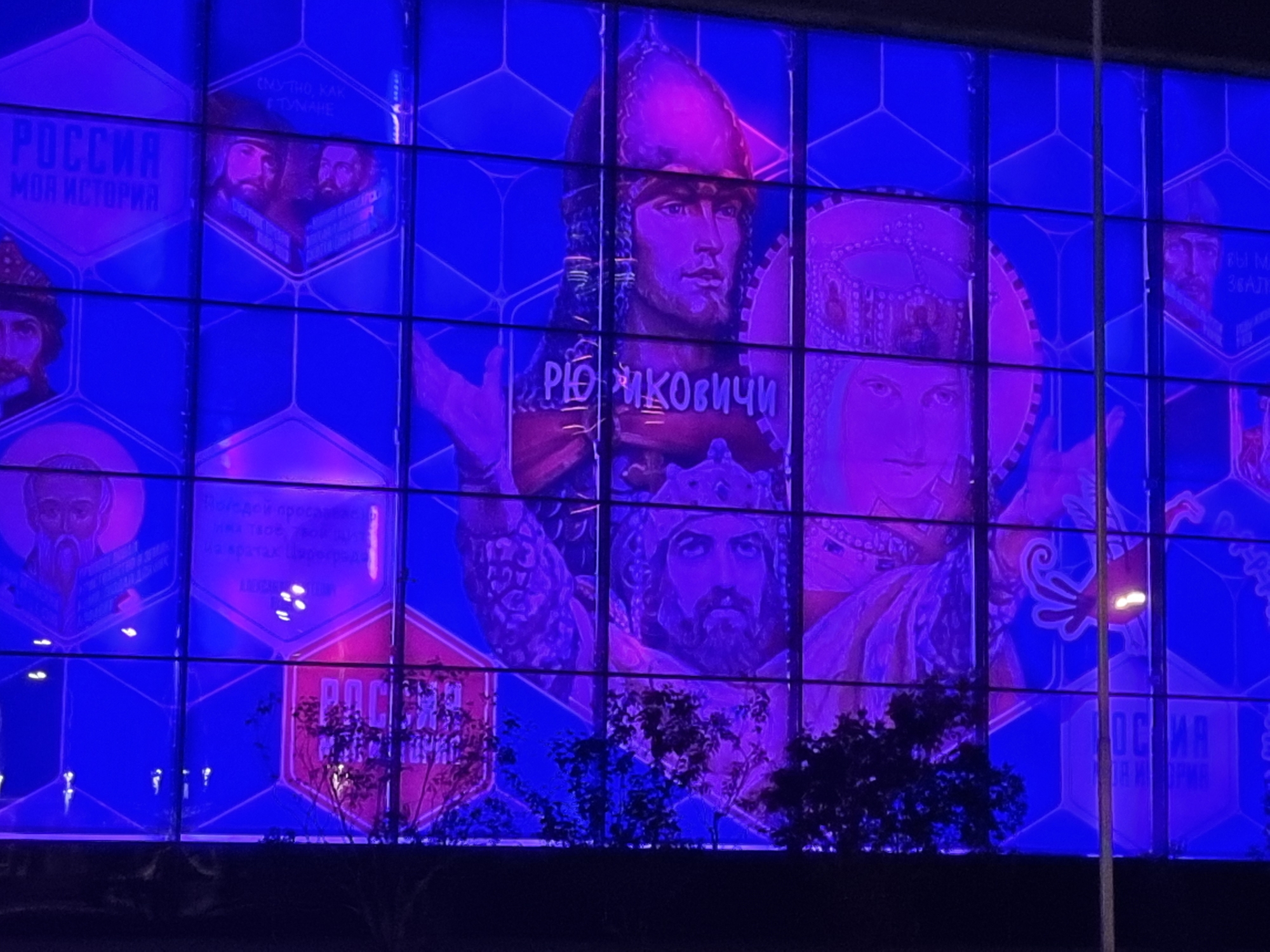 The image shows a large, brightly lit stained-glass window at night, located in Moscow, Russia, specifically at the "Russia. My History" pavilion in VDNKh. The window is divided into hexagonal panels, each depicting historical figures and scenes from Russian history. The dominant colors are vibrant blues and purples, illuminating the artwork and casting a colorful glow on the surrounding area. Silhouettes of trees and bushes are visible in the foreground, suggesting an outdoor setting. The overall impression is one of a grand, illuminated facade, showcasing a patriotic historical narrative.