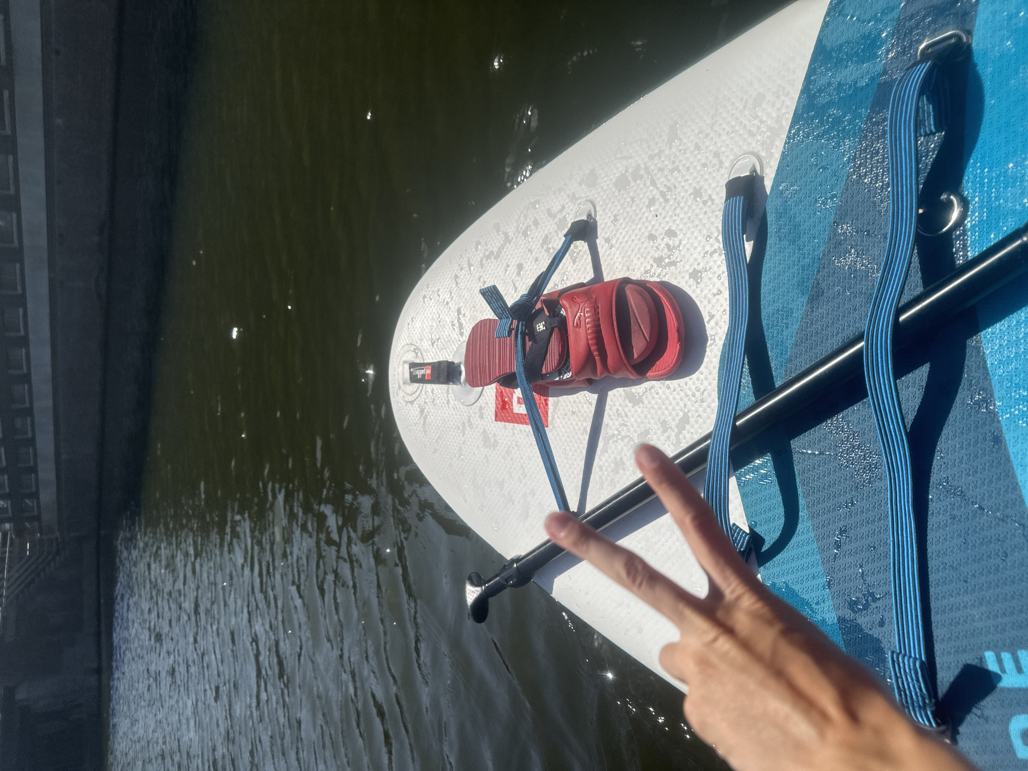 The image captures a first-person perspective from a paddleboard on a body of water, likely a canal in Amsterdam, Netherlands, given the architectural style of the building in the background. The water appears dark and slightly choppy, with sunlight glinting off its surface.
In the foreground, the white surface of the paddleboard is visible, with a red Puma slide sandal secured to it by blue elastic straps. A paddle shaft, dark in color, lies horizontally across the paddleboard. To the left, a hand is positioned, with the index and middle fingers extended in a peace sign. The hand is tanned and appears to be resting on the paddleboard.
The background shows a dark-colored building with multiple windows or rectangular openings, suggesting an urban setting. The presence of the paddleboard and the water indicates an activity like paddleboarding or kayaking in a waterway within the city. The scene is bathed in bright sunlight, suggesting it is daytime.