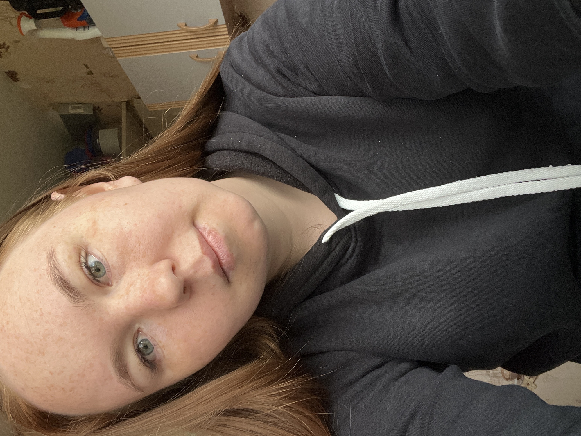 A close-up selfie captures a young individual, likely female, with light brown to auburn hair and distinctive green eyes. Her fair skin is adorned with numerous freckles, particularly visible across her nose, cheeks, and forehead. She has light-colored eyebrows and visible eyelashes. Her lips are slightly parted, and she maintains a neutral to softly contemplative expression while looking directly at the camera.
The person is wearing a black hooded sweatshirt (hoodie) with a textured fabric, and prominent white flat drawstrings are visible near the neckline. Part of her hair falls over her shoulder and chest.
The background, appearing rotated due to the angle of the selfie, shows an indoor setting. Indistinct furniture, possibly a dresser or cabinet with light wooden handles and light gray/brown drawers, is visible. Other cluttered items suggest a personal or domestic space. There are no specific cues to indicate the time of day or weather, but the lighting suggests an indoor environment. No text is visible in the image. The photo's context is Chaikovsky, Russia.