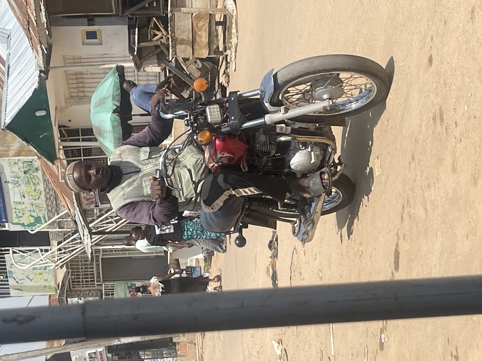 A man wearing a dark long-sleeved shirt, a grey vest, and a cap rides a red and chrome motorcycle on a dirt road in what appears to be a busy marketplace in Abuja, Nigeria. The man has dark skin and is looking forward. The motorcycle has a square headlight and orange turn signals. Behind the man, other people are visible, some walking and some standing. In the background, there are buildings with corrugated metal roofs and some market stalls. An open green umbrella is set up near the buildings. The ground is dusty, with scattered debris.