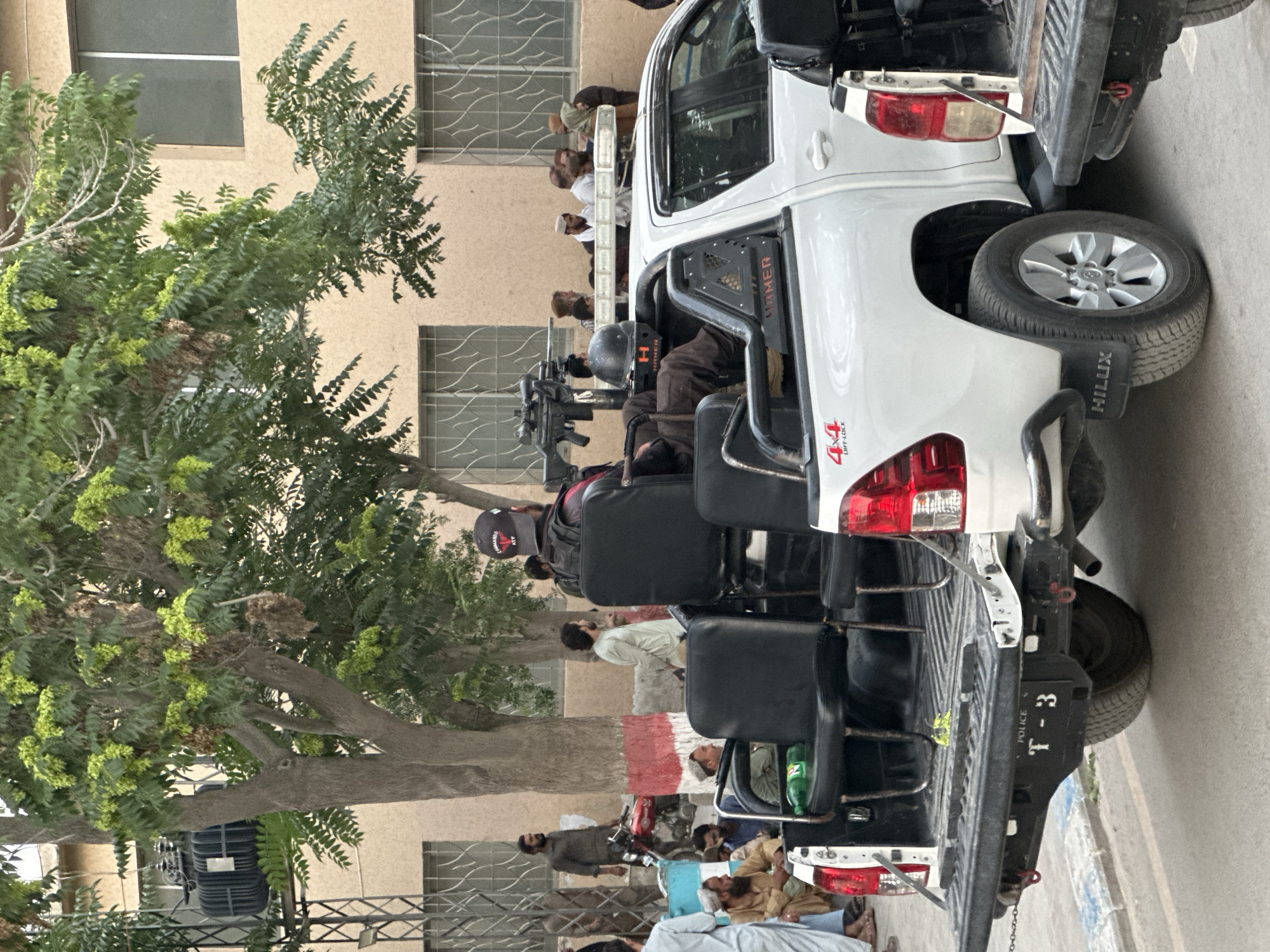 The image depicts a scene in Quetta City Tehsil, Pakistan. A white Toyota Hilux pickup truck, identifiable by the "HILUX" and "4x4" markings, is parked on a paved surface. Two individuals are seated in the bed of the truck, equipped with helmets and what appears to be a heavy weapon. The truck's rear license plate is marked with "POLICE" and "T-3". Several people are present in the background, some standing and some appearing to be resting or sitting on the ground. A large tree with green foliage is visible on the left side of the frame, partially obscuring a building with barred windows. The overall environment suggests an urban setting.