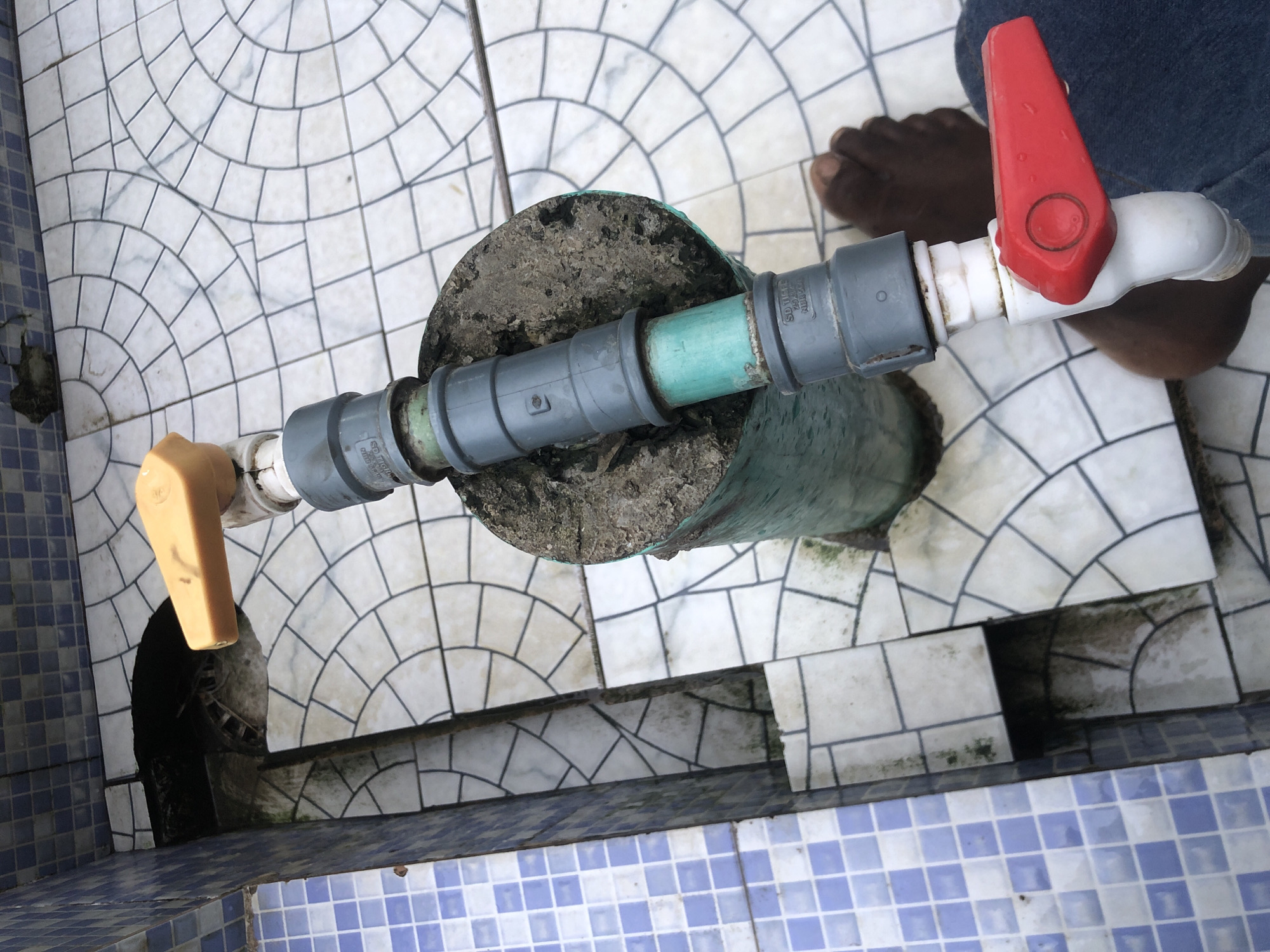 A horizontal outdoor medium shot shows a partially visible person wearing blue denim jeans and dark skin. The person's left foot and lower leg are visible, positioned near the bottom right of the frame. In the center of the image, a PVC pipe assembly extends vertically. The top of the assembly features a yellow handle, likely a valve. Below this, grey PVC pipes are connected with a section of green pipe in between, surrounded by a concrete base. The pipe assembly is situated on a tiled floor. The tiles are white with black grout, arranged in a pattern of concentric arcs and straight lines. To the left of the pipe assembly, a blue and white tiled wall is visible, suggesting an outdoor location, possibly a courtyard or patio. The background shows the continuation of the tiled floor and a dark opening in the ground near the wall. The scene appears to be in Ado, Nigeria, based on the prompt.