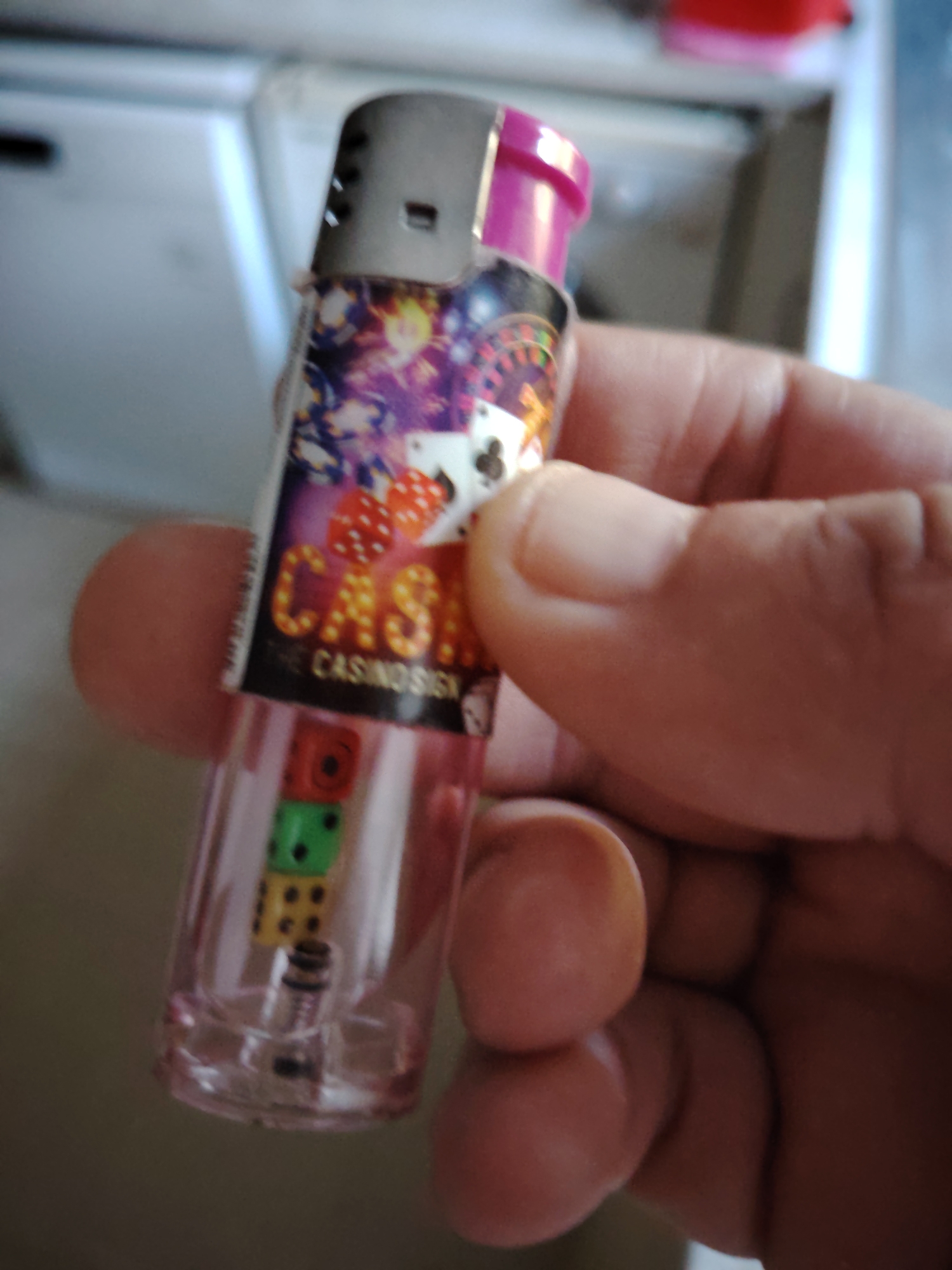 A hand holds a clear plastic lighter with a pink cap. The lighter is decorated with casino-themed imagery, including dice and playing cards. Inside the lighter, three small dice are visible: a red one on top, a green one in the middle, and a yellow one at the bottom. The lighter's wick and flint mechanism are visible at the base. The background is blurred, but appears to be an indoor environment with a white appliance. The image was taken in Huntingdonshire, United Kingdom.