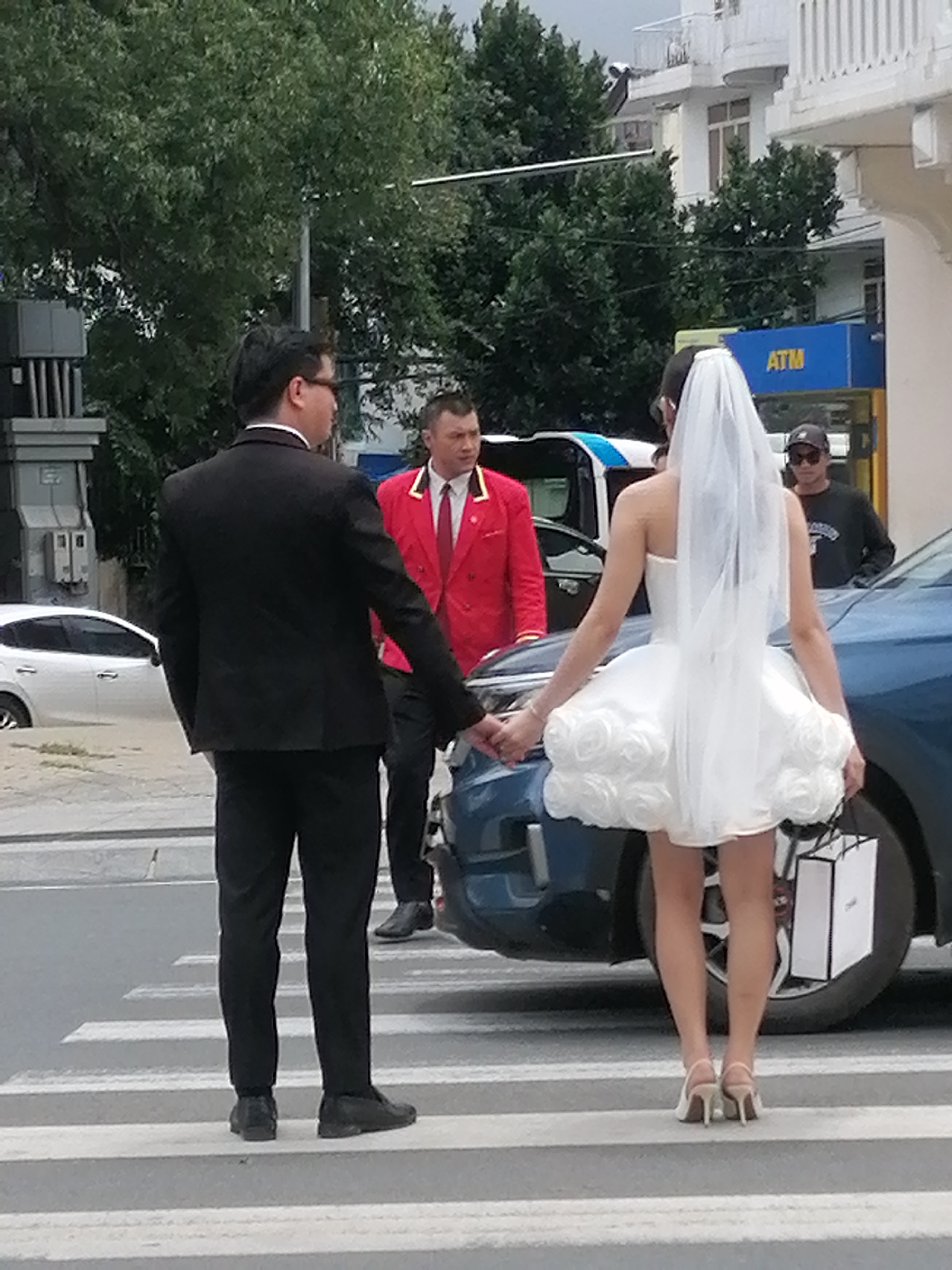 In Da Lat, Vietnam, a wedding couple holds hands while crossing a pedestrian walkway. The groom, dressed in a black suit, stands to the left, while the bride, in a short white wedding dress and veil, is on the right, facing away from the camera. Behind them, a man in a red jacket and a man in a dark shirt are visible. A blue car is parked to the right of the couple. The environment appears to be an urban street with trees and buildings in the background.