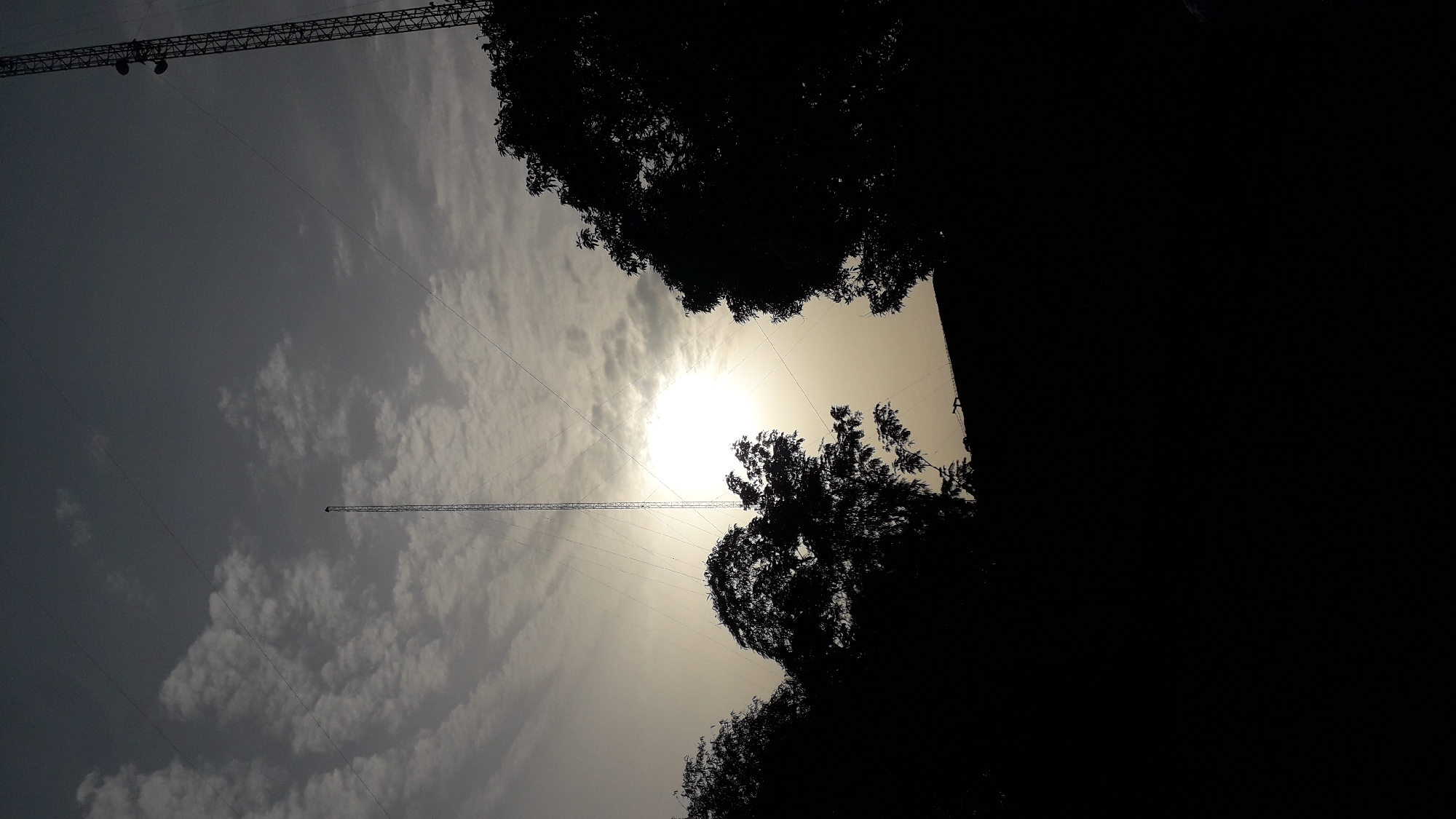 The image shows a view looking up towards the sky, with a bright sun obscured by clouds and trees. Silhouetted tree branches are visible in the foreground, both above and below the sun. Two towers are visible, one towards the upper left and another reaching into the frame from the bottom left. Fine wires connect the towers to the ground. The image is taken during daylight hours. The location is Onitsha, Nigeria.