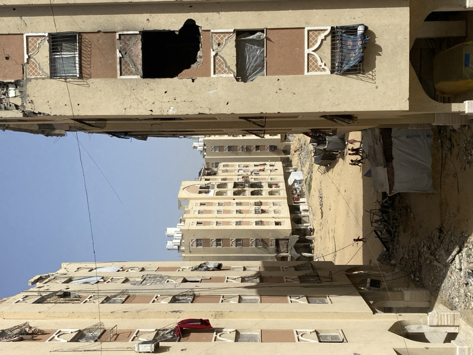 The image depicts a devastated urban residential area in Khan Yunis, Palestinian Territory, characterized by damaged multi-story buildings and an open, sandy ground littered with debris. Two partially visible, light-colored buildings frame the foreground, showing extensive damage including irregular holes, pockmarks consistent with projectiles or shrapnel, and missing sections of facade. Windows on these buildings are either broken, covered with makeshift materials, or feature decorative grilles and arched patterns; a red garment hangs from a window on the left structure. Arched openings are present at the ground level of both foreground buildings, with a yellow object partially visible beneath the right one.
The central ground area is unpaved, sandy earth, containing piles of rubble, construction debris, and refuse. Two makeshift structures, one a white tarp tent displaying a partial UNICEF logo, are located on the right side of this area. Multiple children are present: one child in a red top walks alone towards the left, while a group of approximately five children walks together towards the right. In the mid-ground, a series of similar multi-story residential buildings, also appearing damaged, extend into the background. Many of these background buildings have white cylindrical water tanks on their rooftops. The sky above is clear and light blue, with a few thin wires strung between the foreground structures.