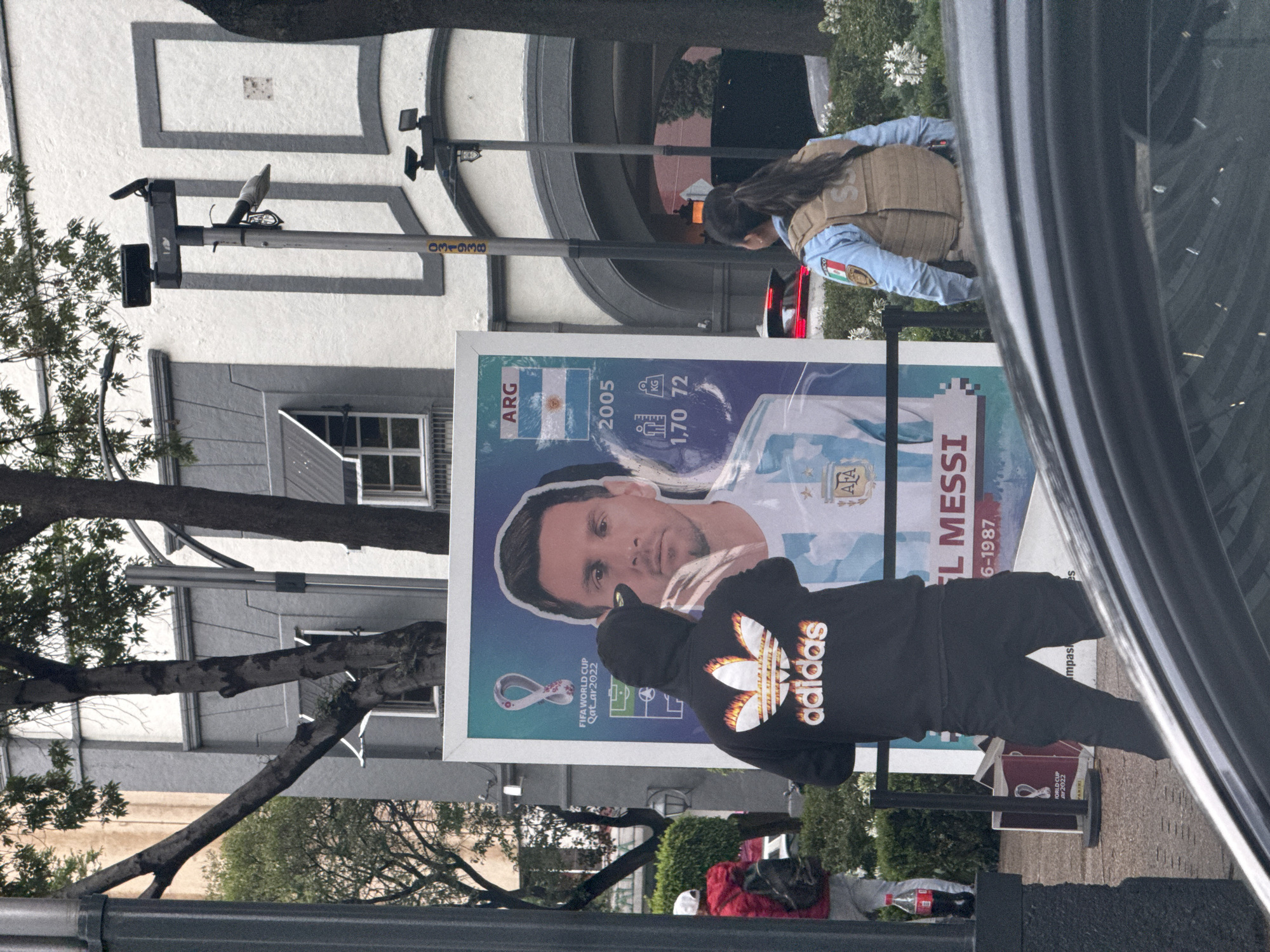 The image shows a poster of Lionel Messi in Mexico City, Mexico. The poster has an Argentine flag, Messi's picture and stats. A person wearing a black Adidas hoodie stands in front of the poster. A female police officer with a protective vest is standing in the background. The number 031938 is visible on the streetlight pole. In the lower portion of the poster, the FIFA World Cup Qatar 2022 logo is displayed. Additionally, two FIFA World Cup Qatar 2022 sticker albums are on the ground.