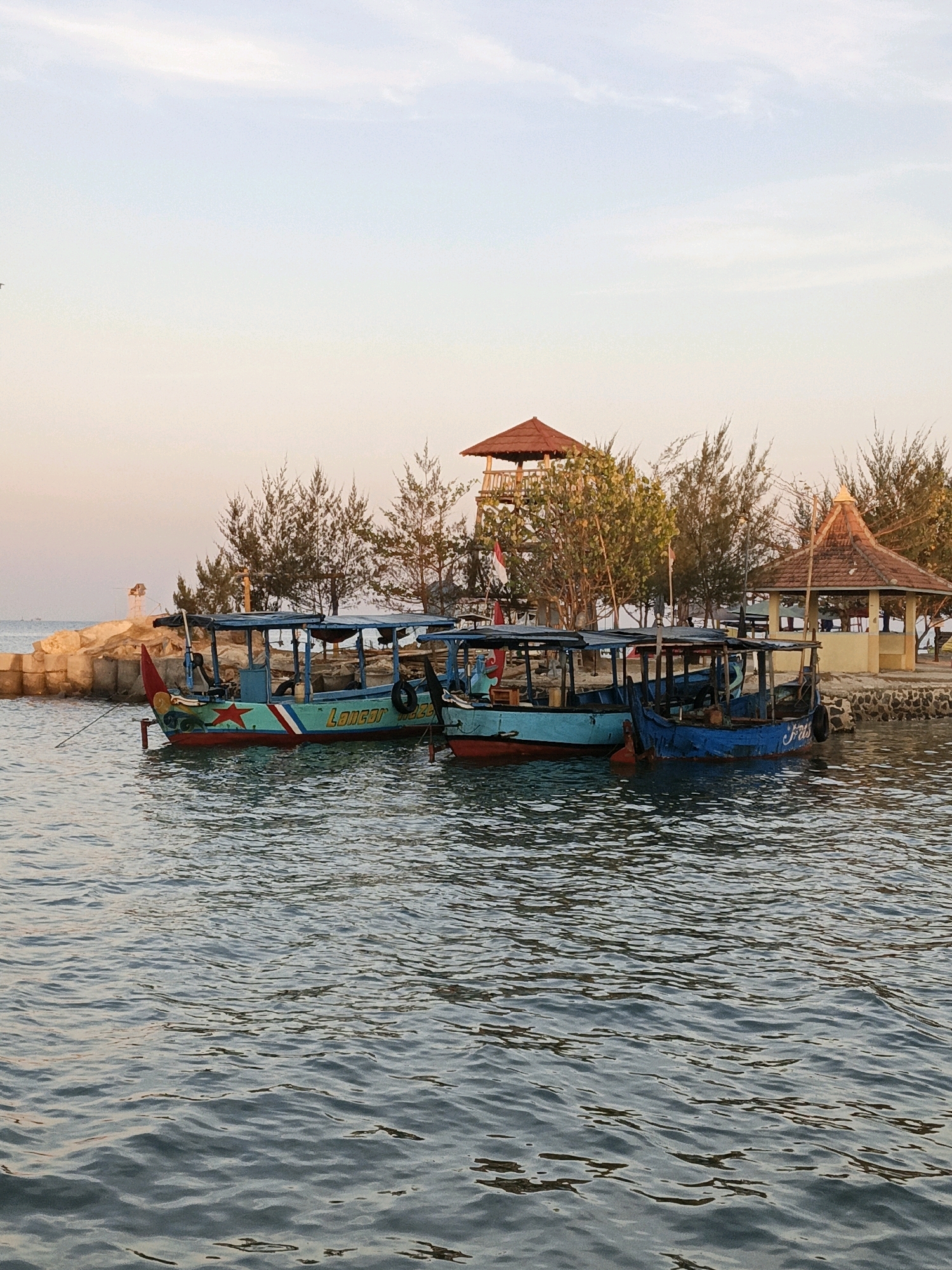 In Cikal, Indonesia, a scenic waterfront is depicted with several boats anchored near a small island. The water is calm, reflecting the soft hues of the sky at what appears to be either sunrise or sunset. The island features a small, reddish-roofed pavilion and a taller observation tower with a similar roof. Several trees with thin, needle-like leaves dot the island's landscape.
In the foreground, three traditional wooden boats are moored side-by-side. The boat on the far left is painted teal and red, with a red star emblem on its side and the word "LANCAR" in green lettering. The middle boat is a light blue color, also with a covered roof structure. The boat on the right is predominantly blue and has the name "FRIS" visible on its side. The boats appear to be waiting for passengers or have recently docked. The background shows a rocky breakwater, and in the far distance, a faint landmass can be seen. The sky is a gradient of soft oranges, pinks, and blues, with wispy clouds. There are no people clearly visible in the image, but the presence of the boats and structures suggests human activity in the area.
