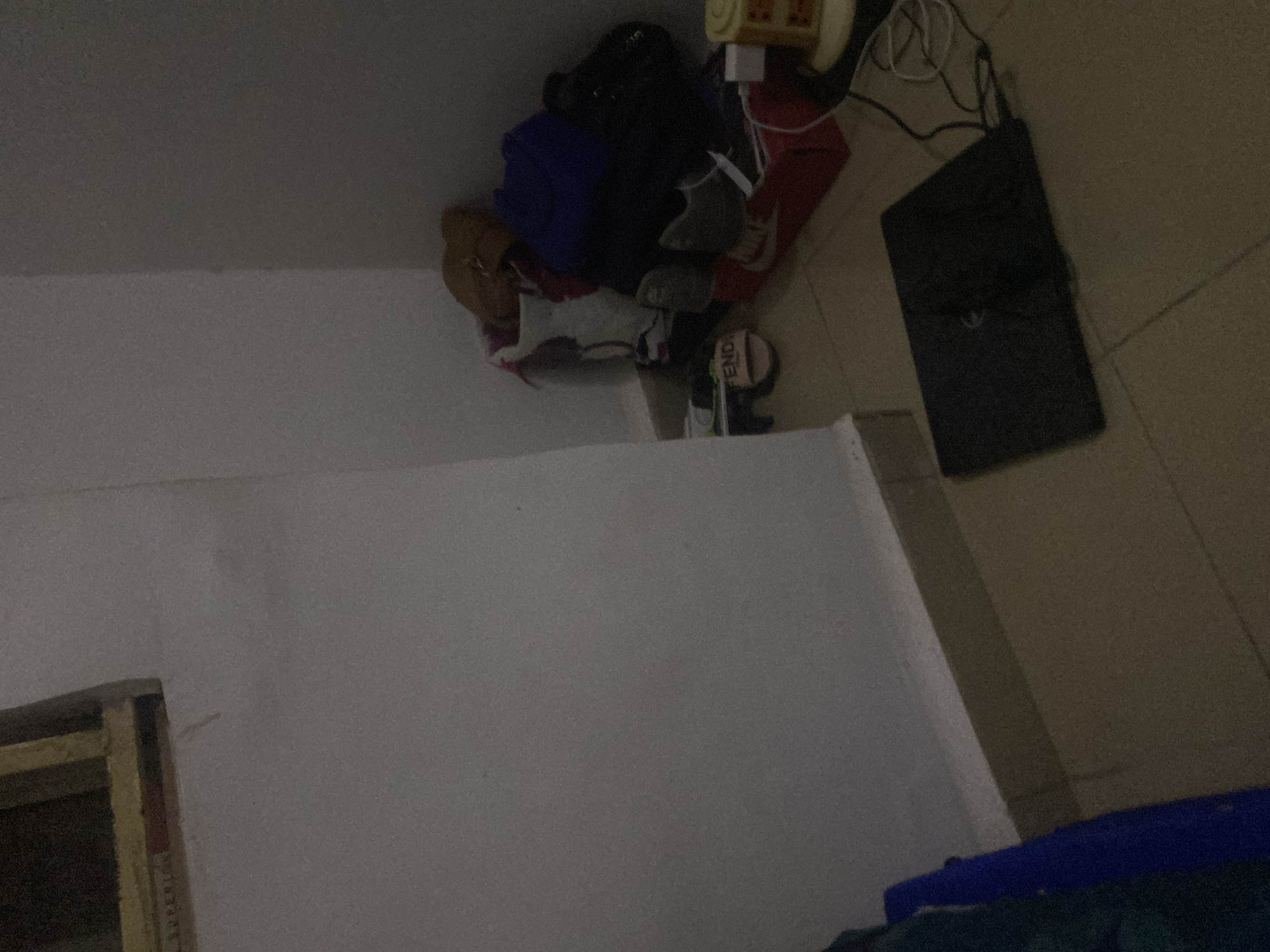 In a dimly lit room, likely a storage area or bedroom in Howayi, Nigeria, various items are piled in a corner. A black laptop is placed open on a tiled floor. Adjacent to it, a red shoebox with white Nike branding is visible, along with other footwear and bags. A pair of light-colored slides with "FENDI" written on them rests on the floor. The walls are painted a light color, and the floor appears to be tiled. The overall impression is one of disarray and clutter.