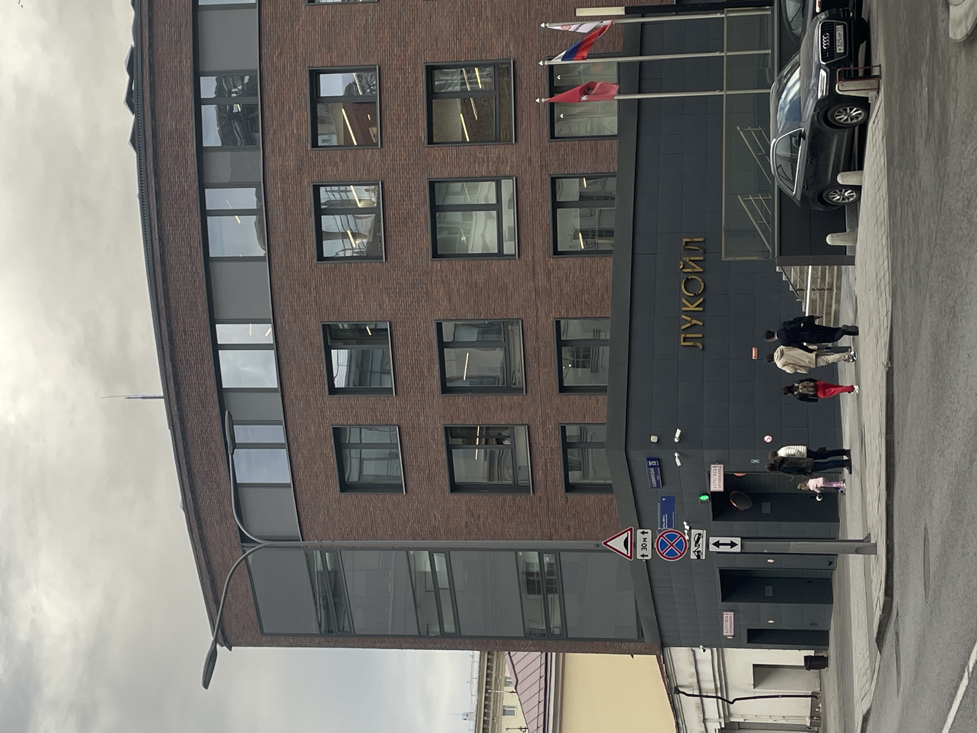 The image shows the front of a building identified as the Lukoil company building in Moscow, Russia. The building has a brick facade with multiple windows and a dark gray section at the entrance displaying the company name in gold Cyrillic letters ("ЛУКОЙЛ"). Russian flags are visible above the entrance. Several people are walking on the sidewalk in front of the building. A black Audi car is parked to the right of the entrance. A street sign with parking restrictions and other road markings is located on the sidewalk. The sky is overcast, suggesting a cloudy day.