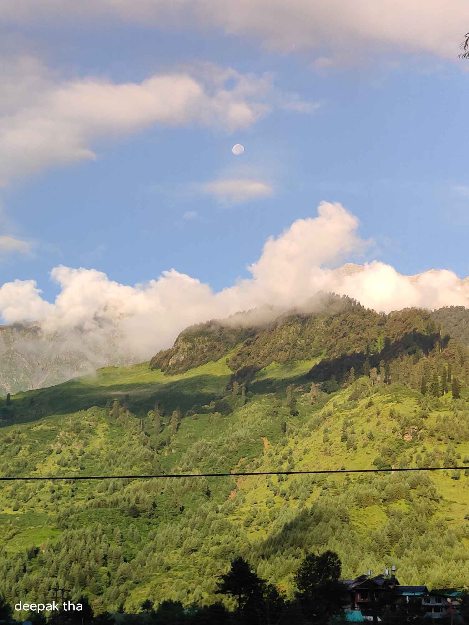 The image captures a scenic mountain landscape in Palchan, India, under a partly cloudy sky. The sun is casting shadows across the green, forested slopes of the mountains. In the distance, the moon is visible, small and white against the blue sky. Some clouds are gathered around the mountain peaks, obscuring parts of them. The foreground shows a dense forest with various trees. A power line stretches horizontally across the lower part of the image. At the very bottom, there are glimpses of houses and structures nestled within the trees. The text "deepak tha" is present in the lower left corner of the image.