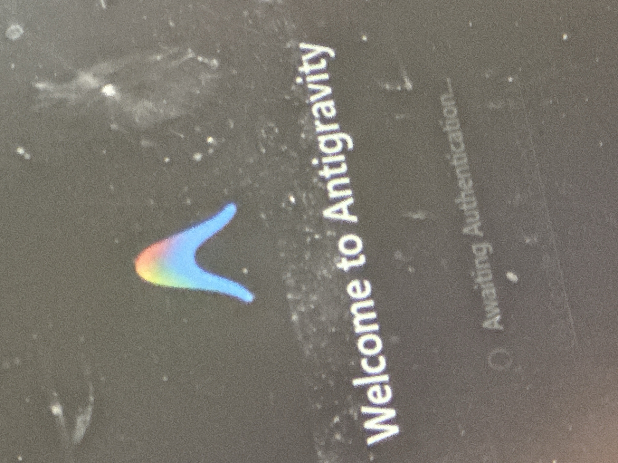 The image shows a screen displaying the words "Welcome to Antigravity" above a V-shaped logo with a rainbow color gradient. To the right, the text "Awaiting Authentication..." is visible. The screen is smudged with dust and fingerprints. The location is unknown and cannot be determined from the provided image. There are no people, actions, or interactions visible.