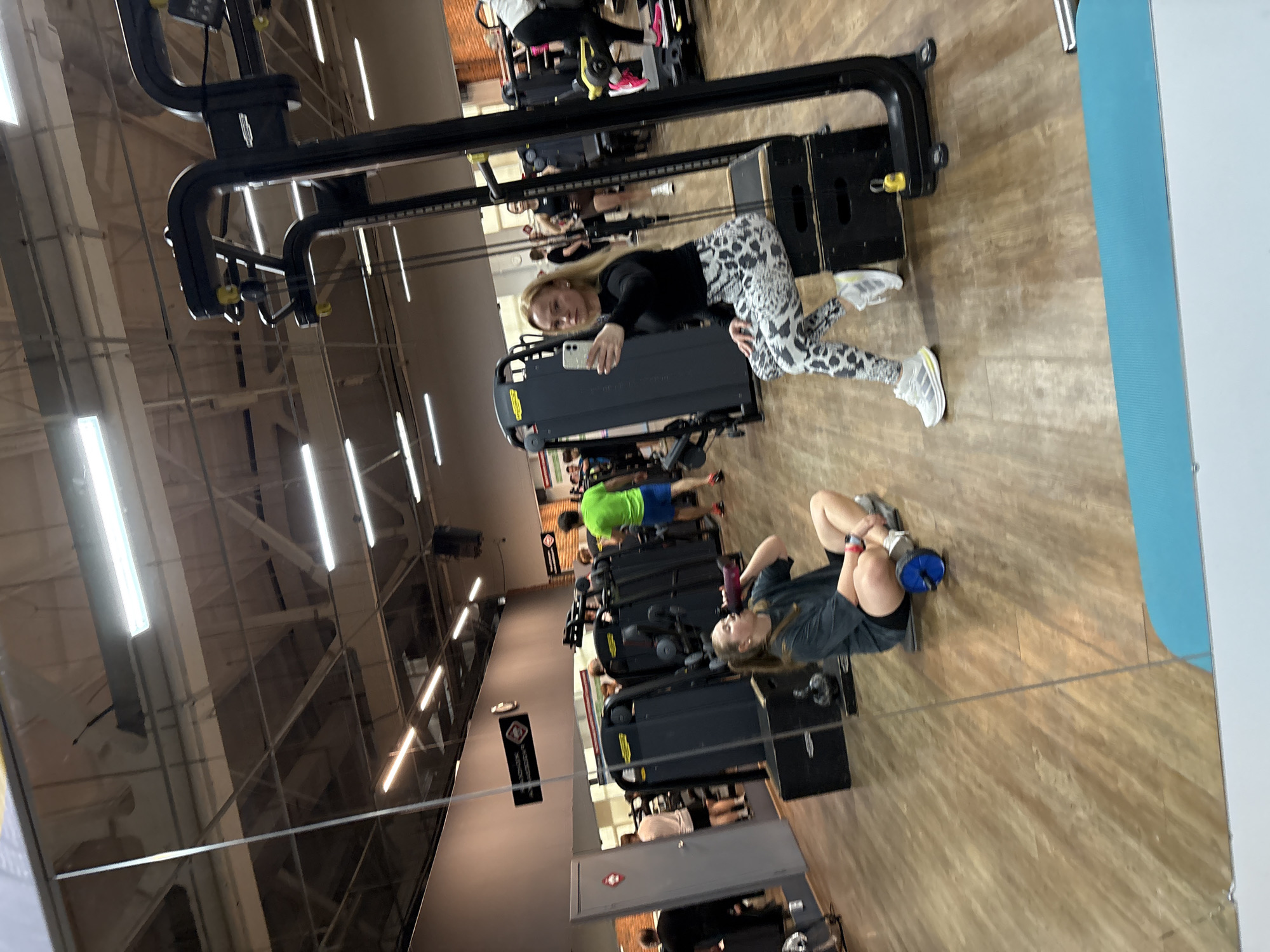 A mirrored image shows a gym interior. The reflection includes various gym machines, individuals exercising, and a person posing for a selfie. The gym has wooden flooring and reflective panels on the ceiling, with fluorescent lighting. The location appears to be in the city of Chaikovsky, Russia.