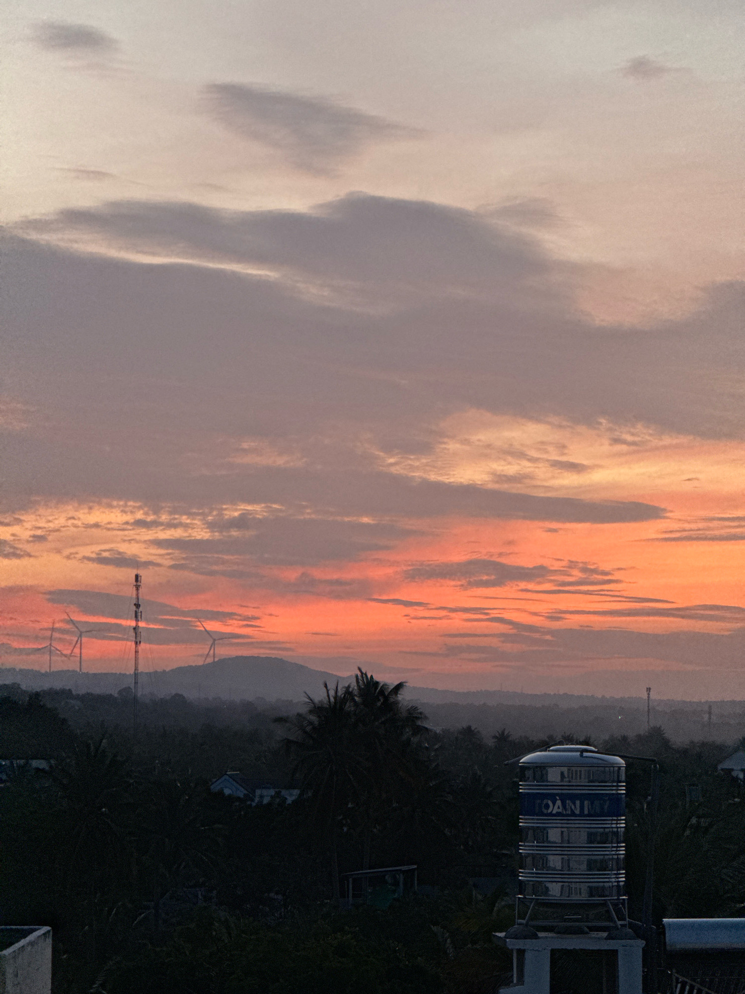 The image depicts a serene sunset over Mũi Né, Vietnam. The sky is painted with soft hues of orange, pink, and purple, transitioning into a pale lavender at the top. Wispy clouds are scattered across the horizon, some catching the last rays of sunlight. In the distance, rolling hills are silhouetted against the vibrant sky. A few wind turbines are visible on the hillsides, hinting at the wind-swept nature of the region.
In the foreground, lush green vegetation, including palm trees, creates a dense canopy. A tall telecommunications tower stands prominently on the left side of the frame. To the right, a large, cylindrical water tank, emblazoned with the blue text "TOÀN MỸ" (meaning "Perfect" or "Entirely Beautiful" in Vietnamese), is mounted on a white pedestal. The tank reflects the dusky light. Several low-lying buildings are nestled within the foliage, their details obscured by the fading light. The overall atmosphere is tranquil and evokes the peaceful end of a day in this coastal town.