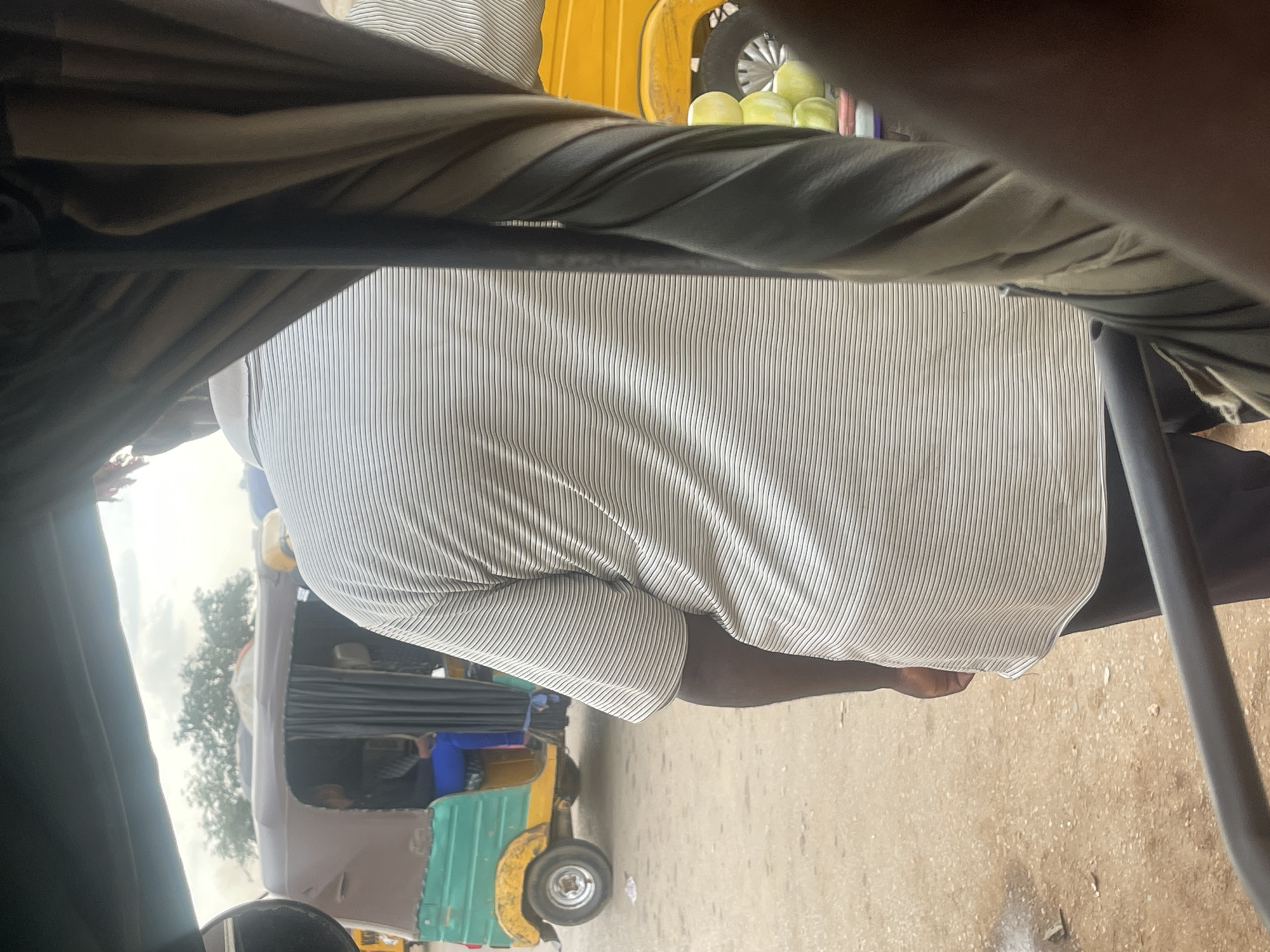 The image shows a man from behind wearing a horizontally striped polo shirt. He is standing on a dirt road in what appears to be a market area. To his left, there is a three-wheeled vehicle, commonly known as a "keke," painted green and yellow, with a person seated inside wearing blue pants. In the background, there are trees and a cloudy sky. To the right of the man, a portion of a yellow vehicle is visible, and some round, light-colored objects, possibly fruits or vegetables, are stacked. The overall environment suggests a bustling, outdoor marketplace.